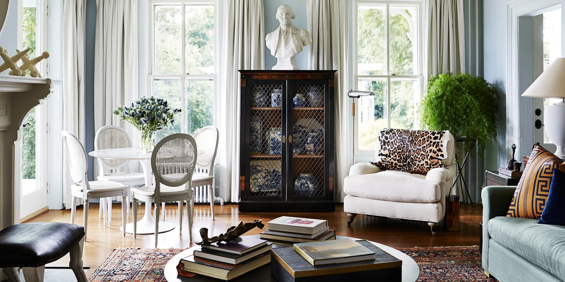 A light blue living room features a black display cabinet filled with china, a white tulip dining table with cane chairs, and a cream armchair.