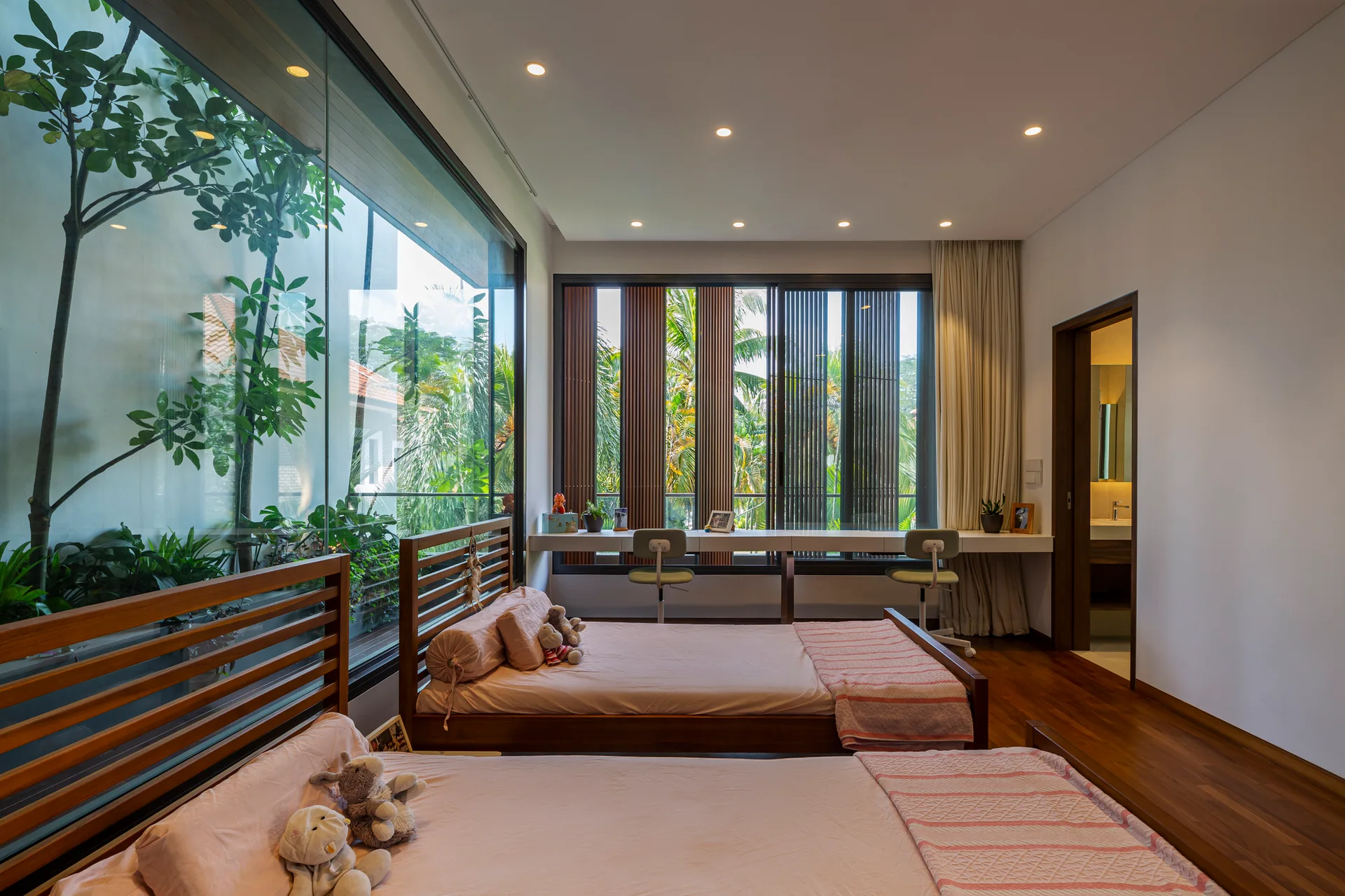 A shared children's bedroom featuring two wooden twin beds, a long white built-in desk beneath a window with vertical slats, and hardwood floors.