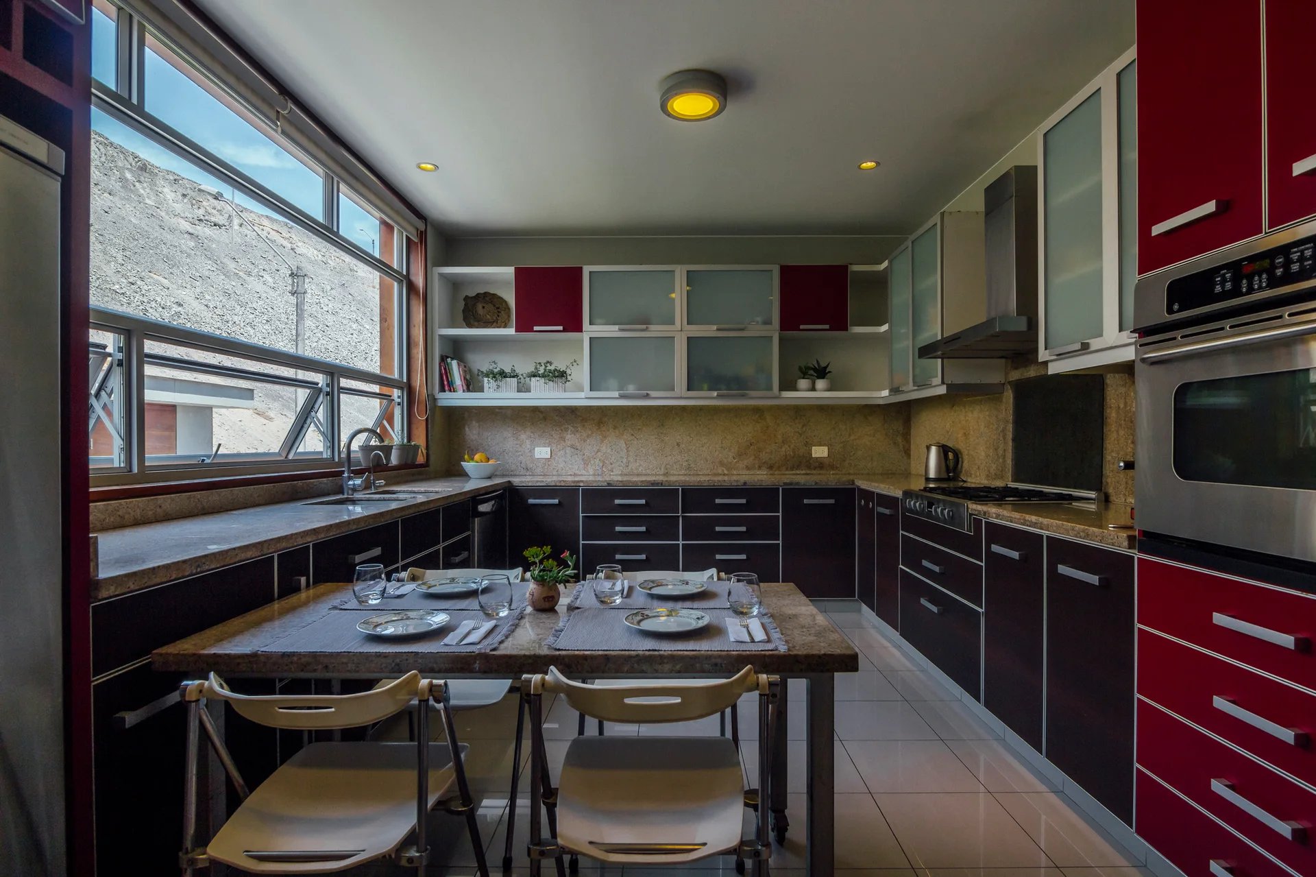 A modern U-shaped kitchen featuring red upper cabinets, dark base cabinetry, a stone-topped dining table with chairs, and a large horizontal window.