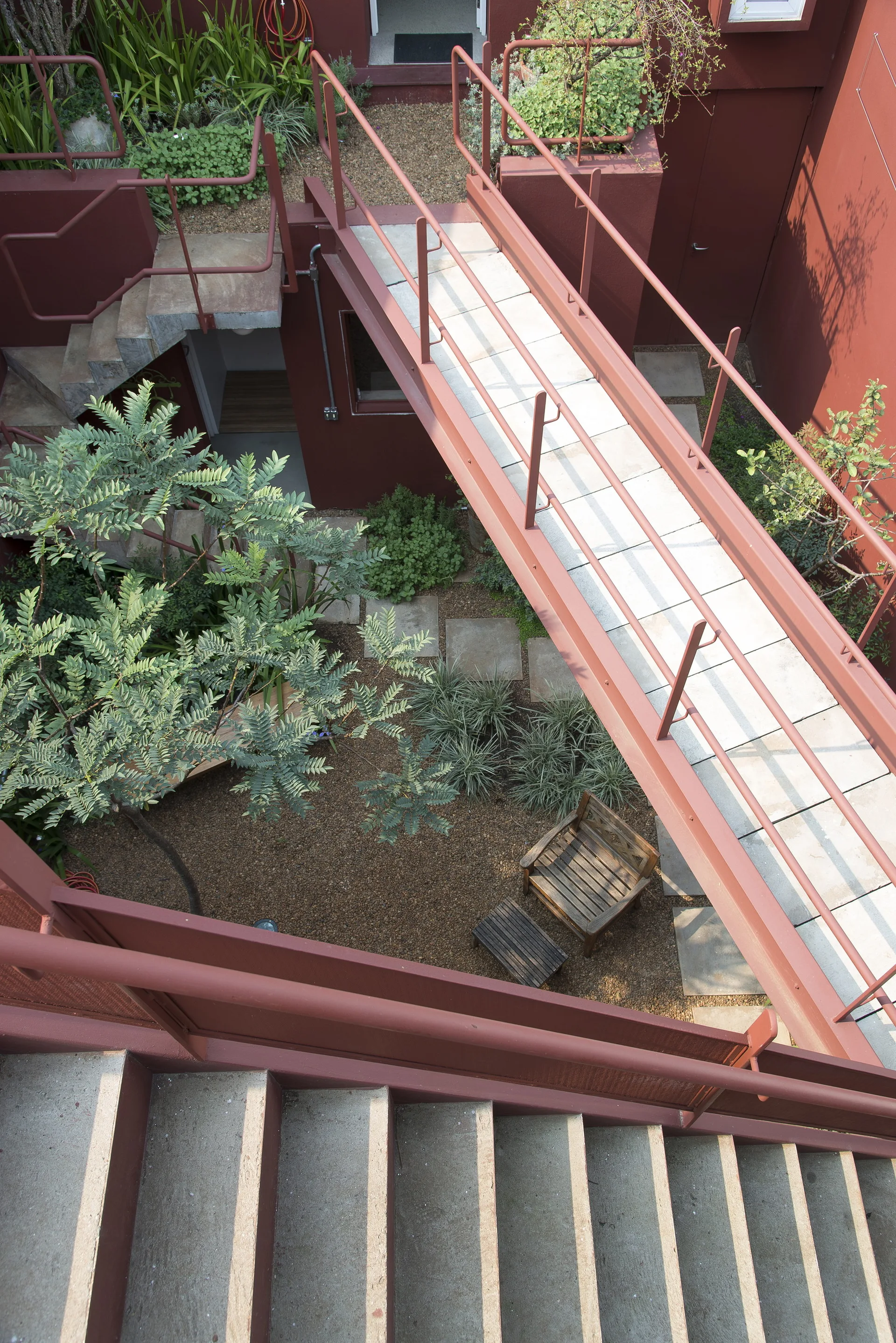 An outdoor courtyard with deep red walls features concrete staircases, an elevated concrete walkway with red railings, and gravel ground cover.