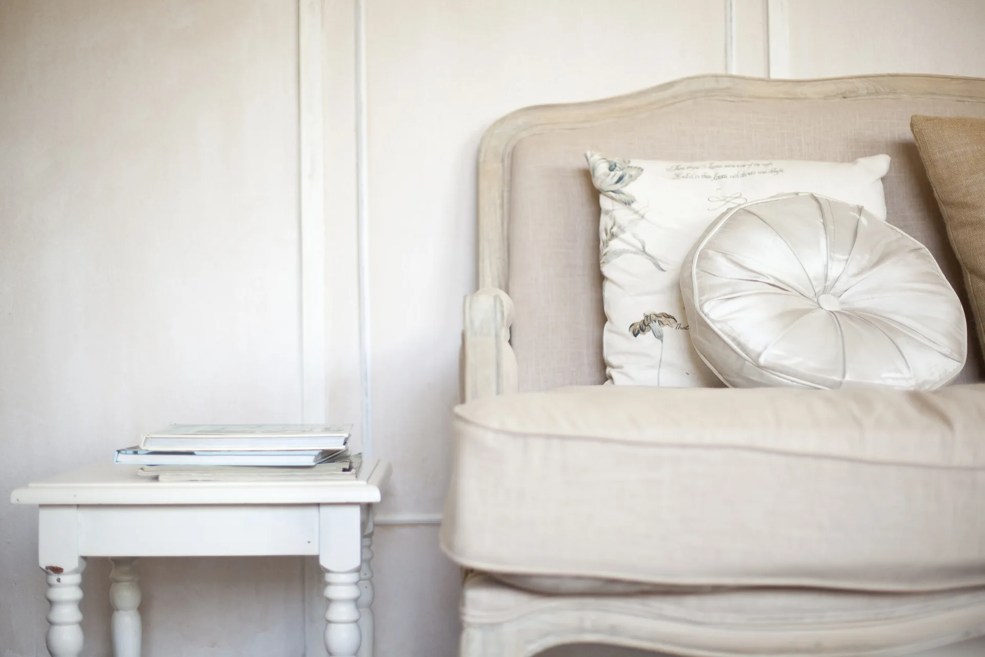 A light beige upholstered armchair with patterned pillows sits next to a small white wooden side table with books against an off-white paneled wall.