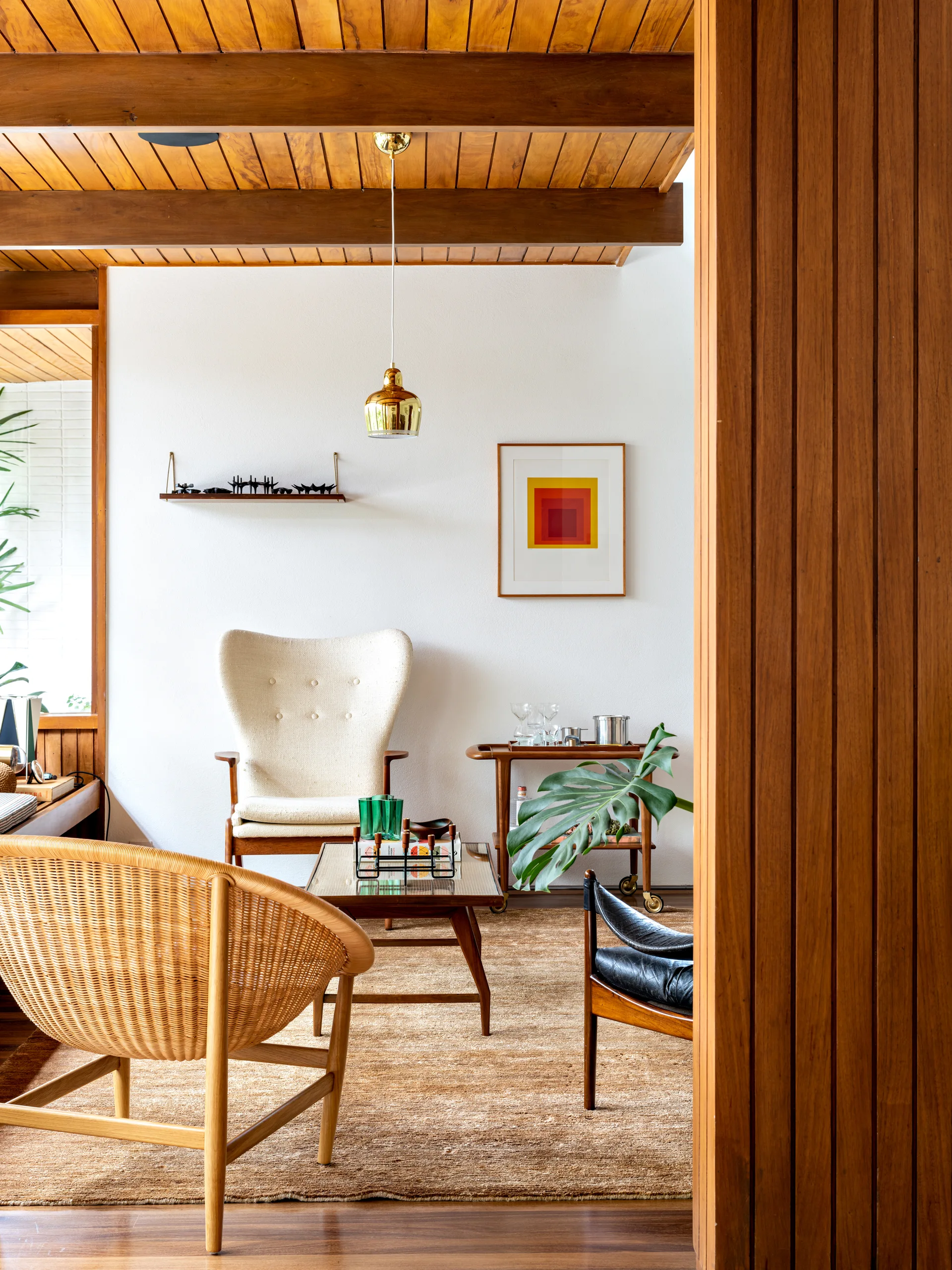 A mid-century modern living room features a wood plank ceiling with beams, a cream wingback chair, a wicker armchair, and a natural fiber rug.