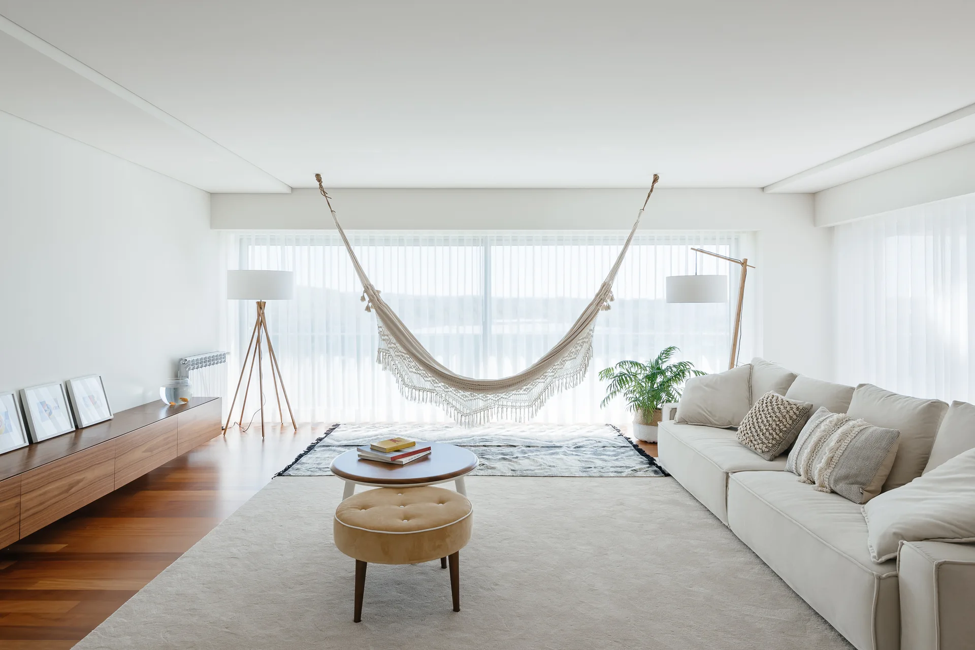 A bright living room features a cream-colored sectional sofa, a hanging fringed hammock by a large window, and a low wooden media console.