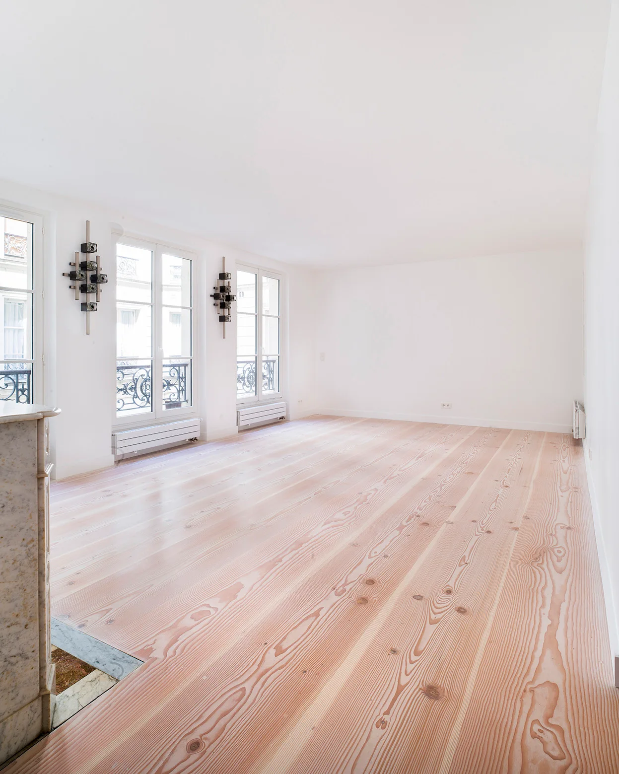 An empty white room features wide-plank light wood flooring with a prominent grain pattern, tall casement windows, and two dark geometric wall sconces.