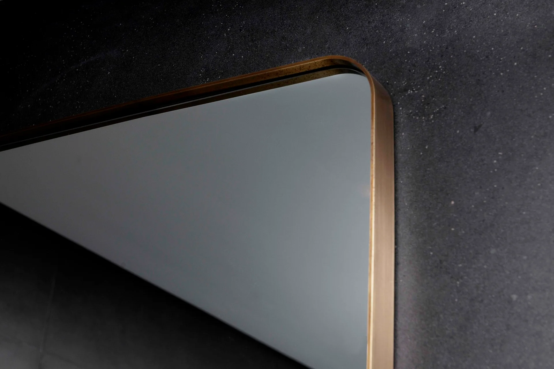 A close-up of a mirror featuring a rounded corner and a brushed brass frame mounted on a dark textured wall.