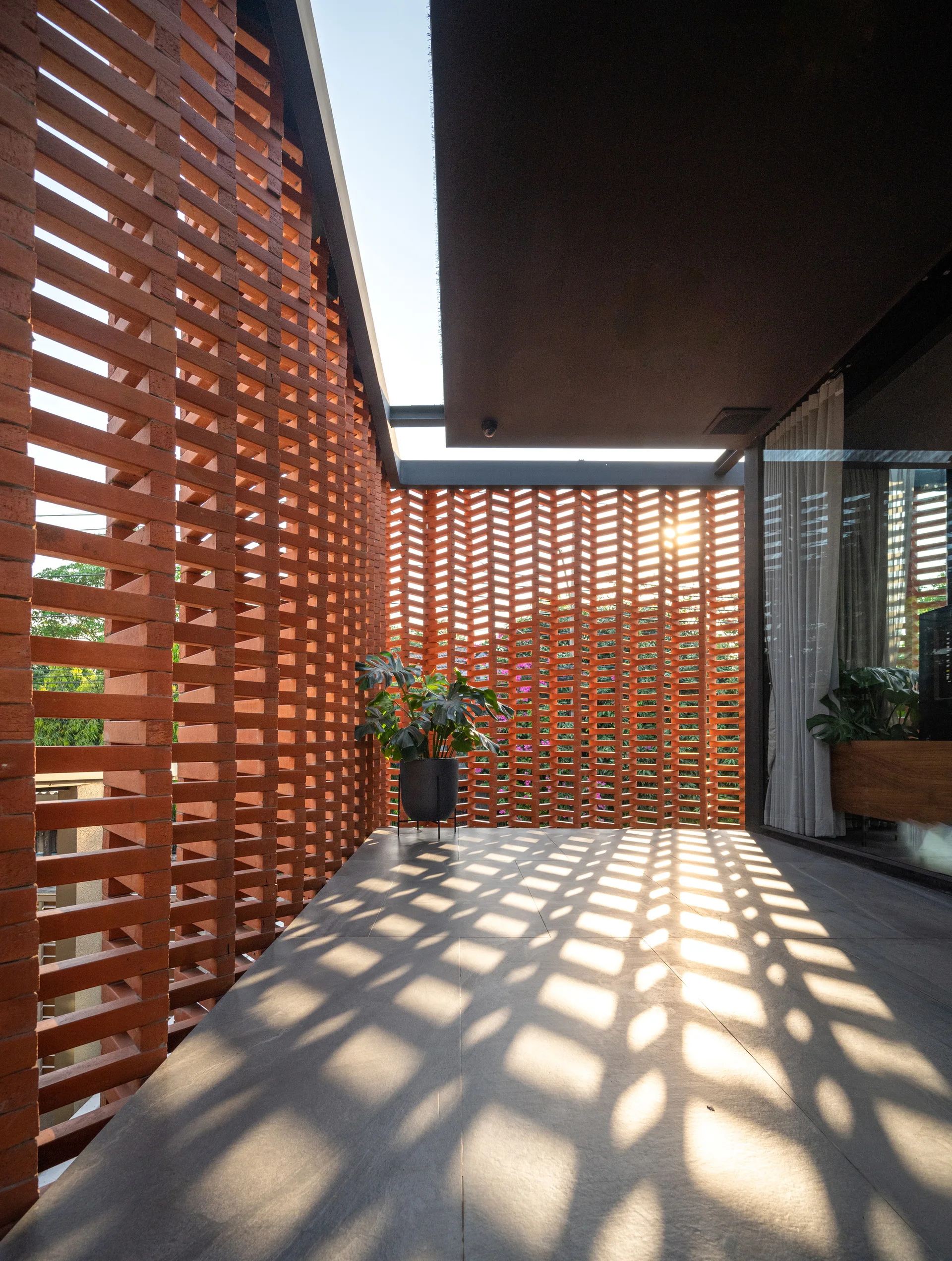 A covered outdoor patio with a red brick screen wall casting grid-like shadows onto a grey tiled floor, featuring a potted plant.