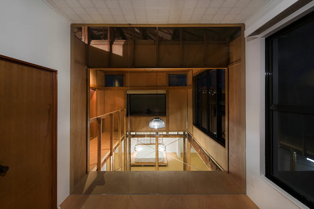 A view from an upper plywood-clad floor looks through a glass partition down to a lower-level bedroom with a platform bed and a hanging pendant light.