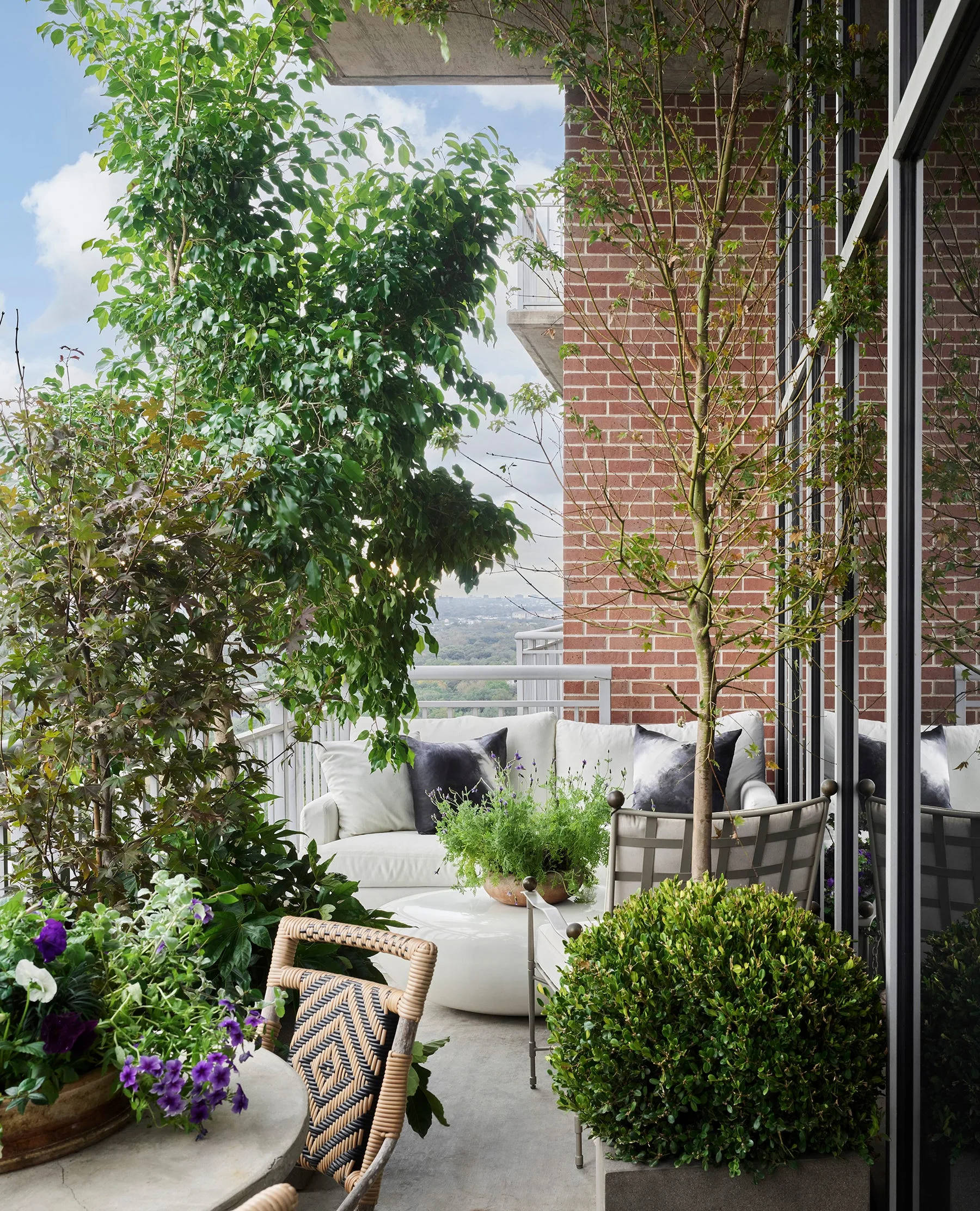 A high-rise balcony features a white outdoor sofa with dark pillows, surrounded by dense green potted plants, a woven rattan chair, and a brick wall.
