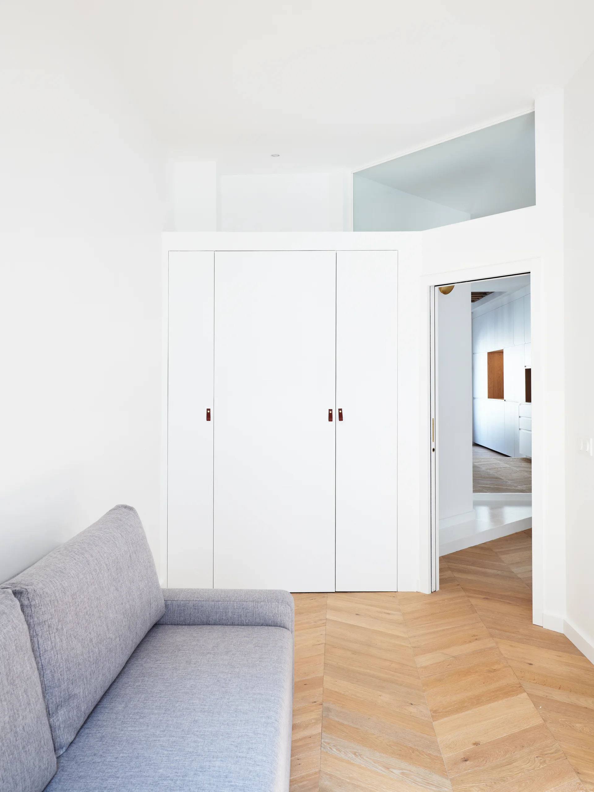 A minimalist room featuring a grey sofa, a tall white wardrobe with leather pull tabs, and light wood chevron parquet flooring.