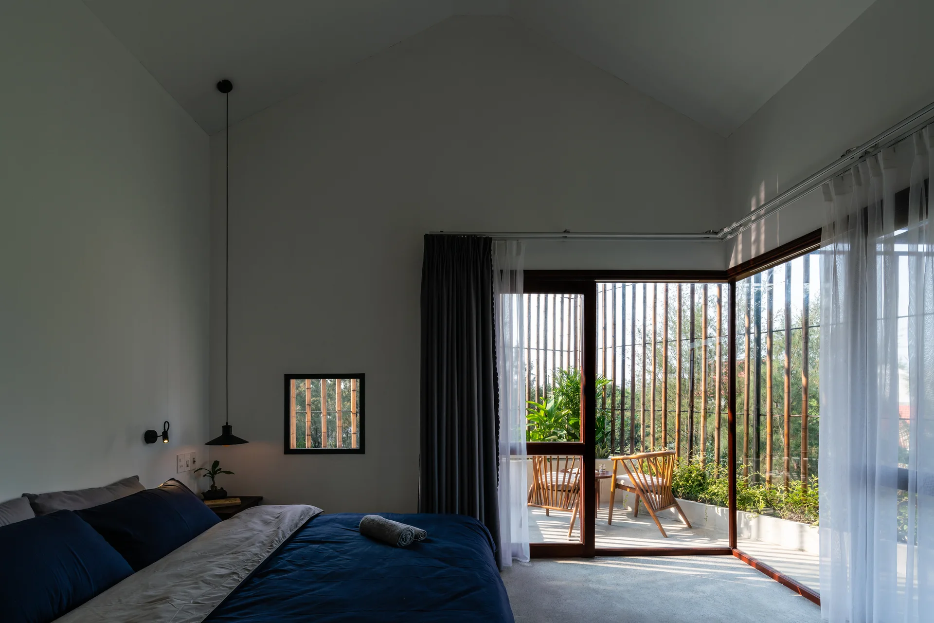 A minimalist bedroom with white walls, navy blue bedding, and a corner glass wall opening to a balcony with vertical bamboo screens.