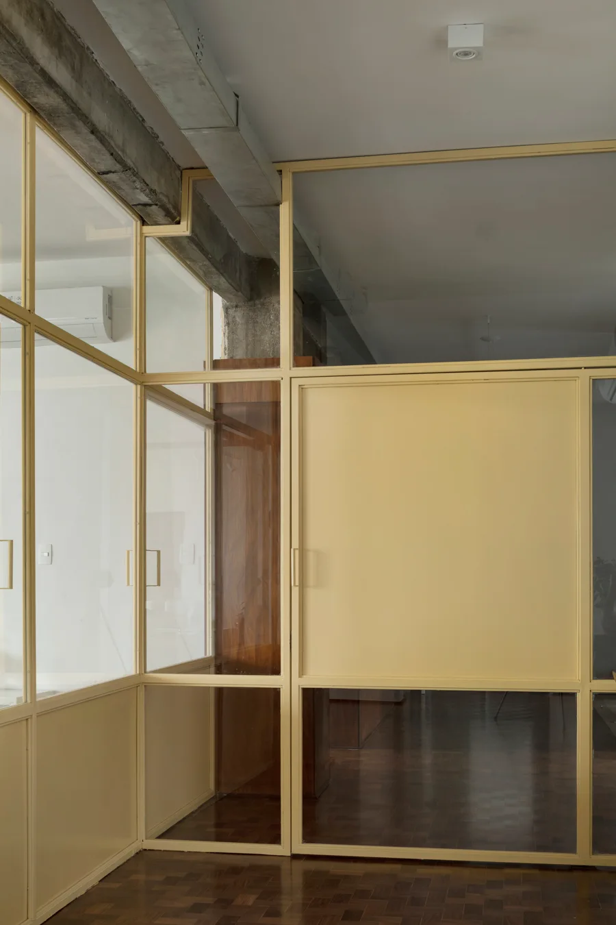 A yellow metal and glass partition wall divides an interior space featuring dark wood parquet floors and an exposed concrete ceiling beam.