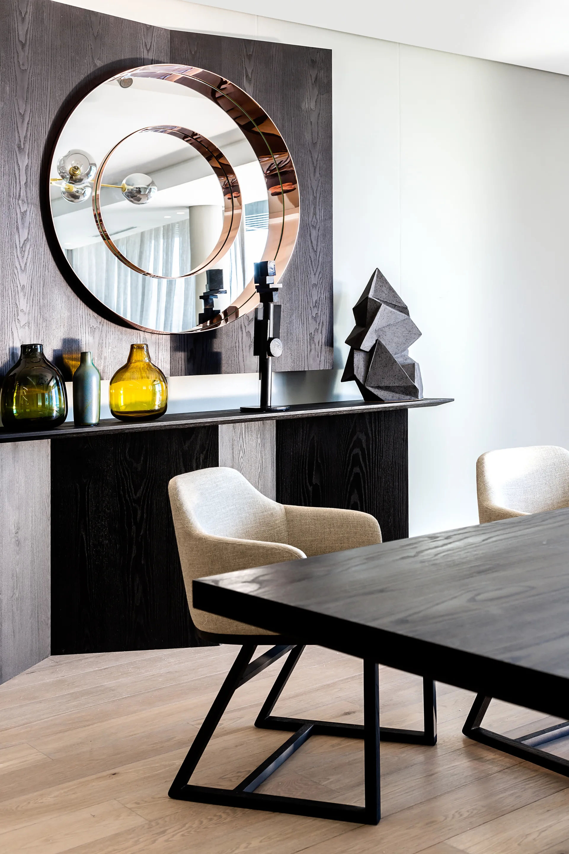 A dark wood console with a circular copper-framed mirror holds glass vases and sits behind upholstered dining chairs in a modern dining space.
