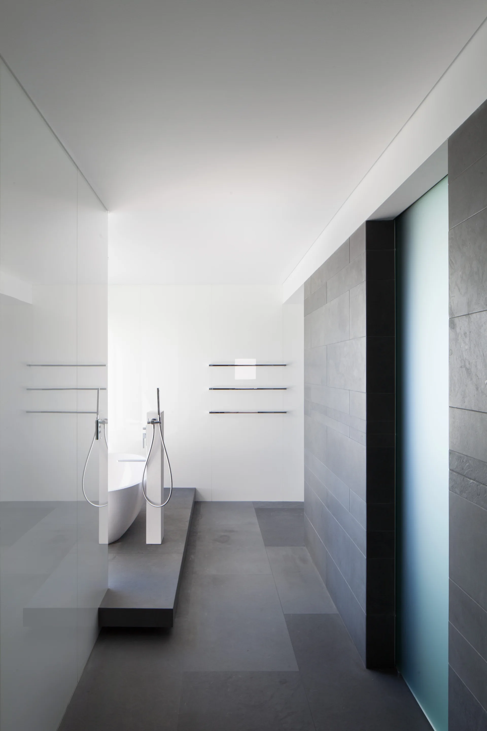 A white freestanding bathtub sits on a raised dark grey tile platform in a minimalist bathroom with high-contrast grey and white walls.