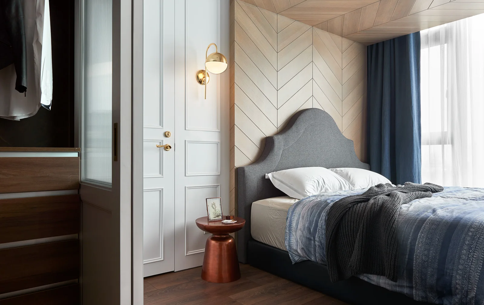 A bedroom features a grey upholstered headboard set against a light wood herringbone wall, a metallic copper nightstand, and a white paneled door.