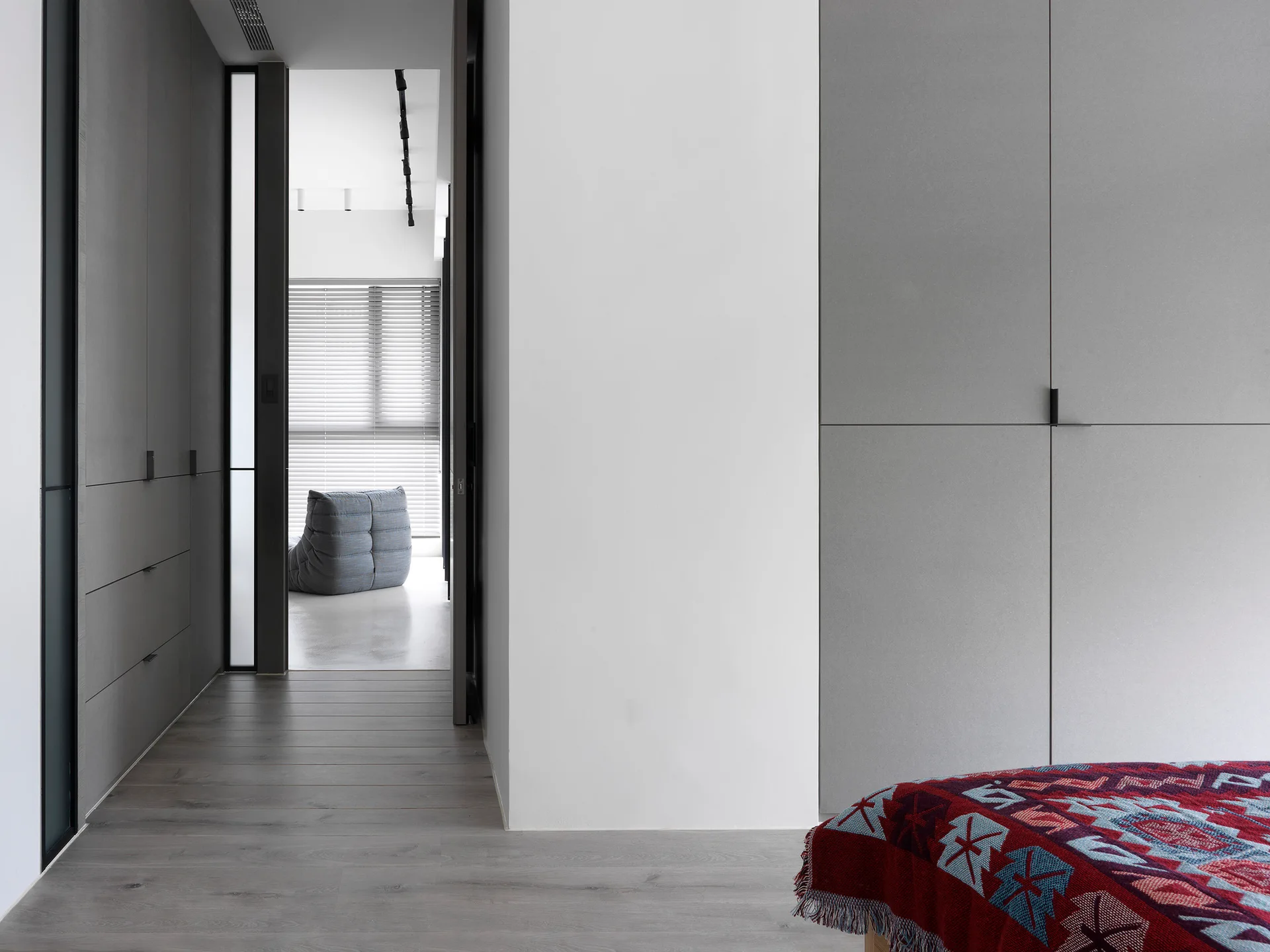 A minimalist bedroom featuring matte grey built-in wardrobes, grey flooring, and a patterned red blanket with a view into an adjacent sunlit room.