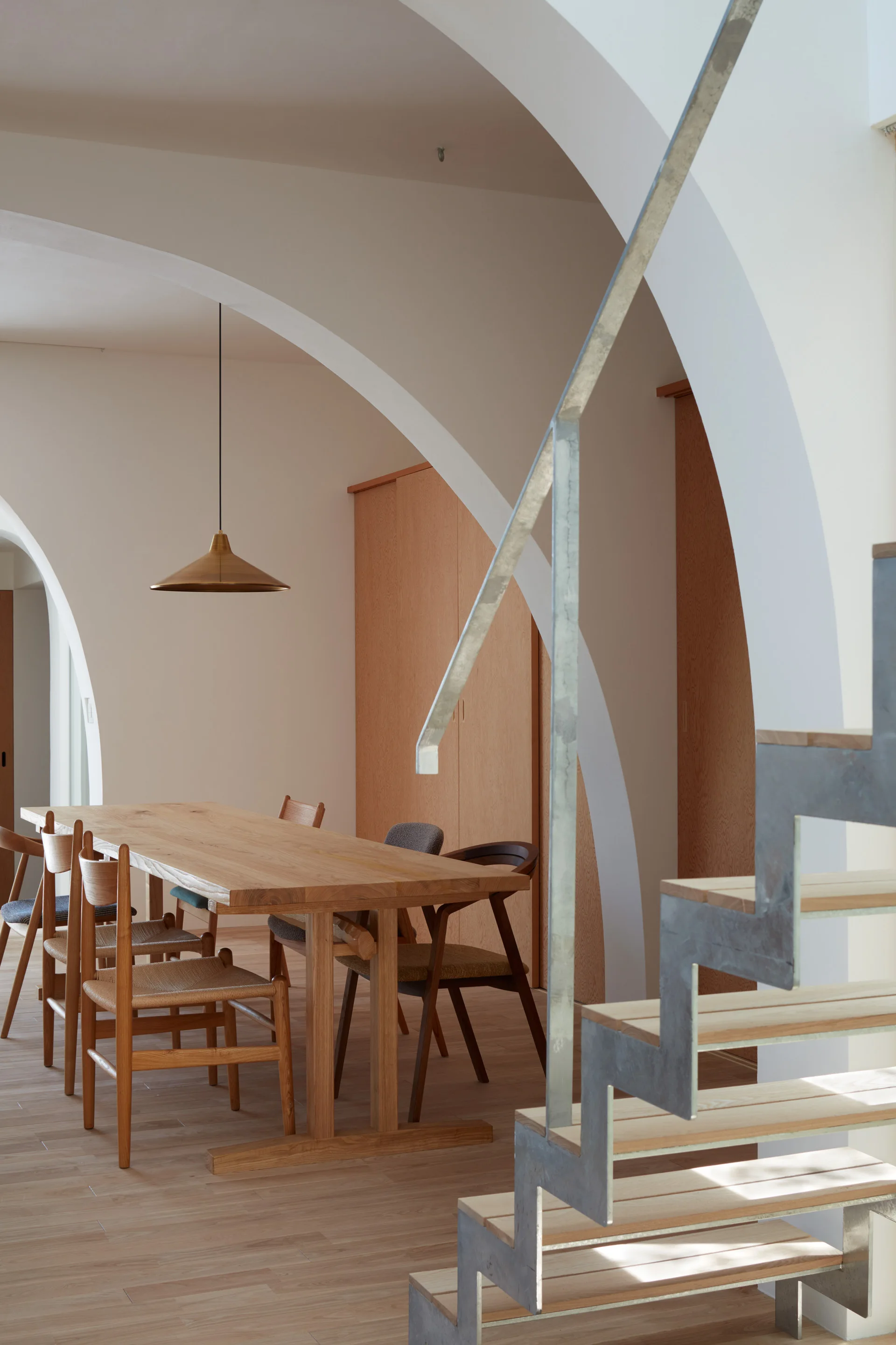 A minimalist dining room with a light wood table and chairs framed by white plaster archways next to a metal staircase.