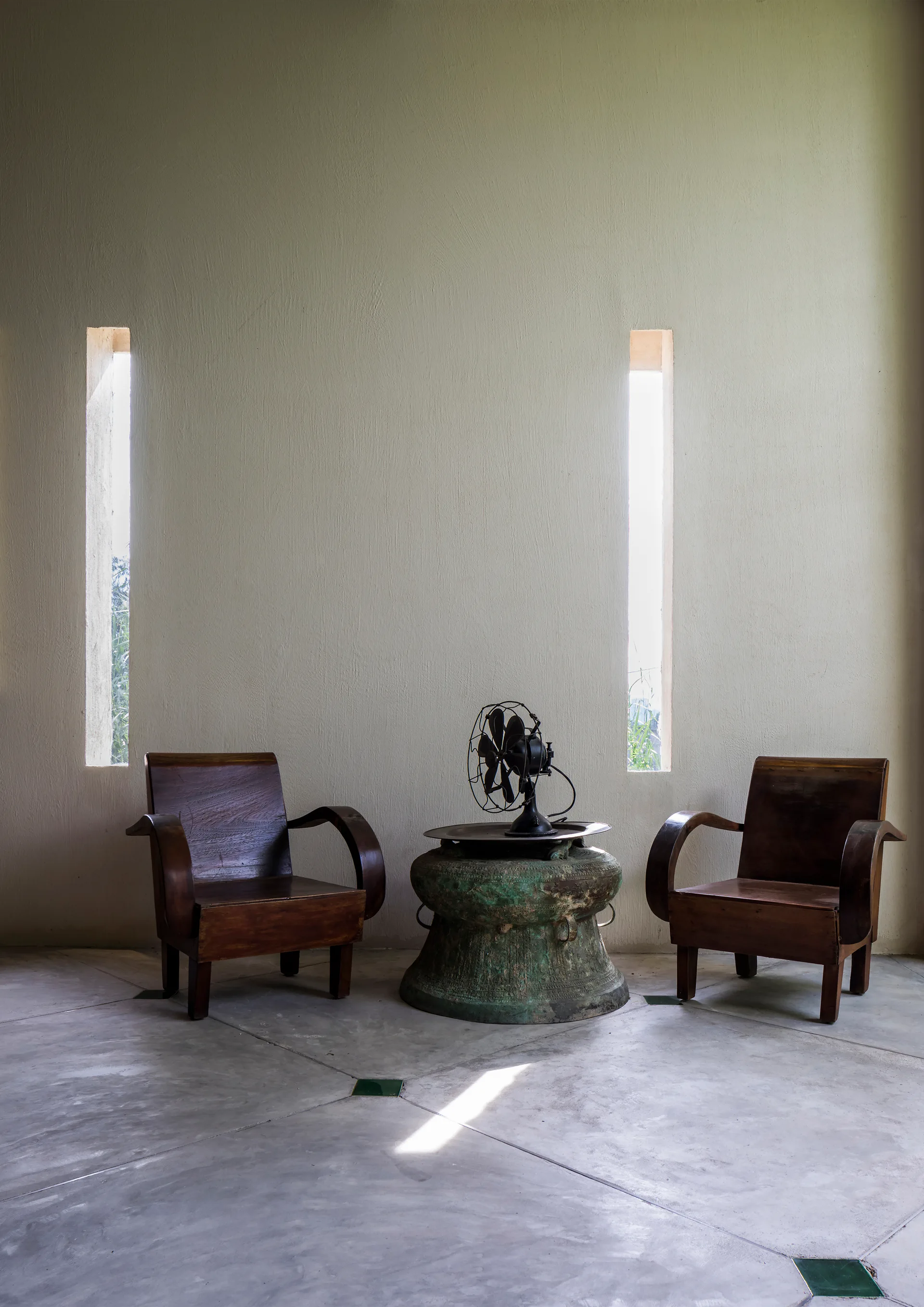 Two wooden armchairs flank a weathered metal table with a vintage fan, set against a wall with narrow vertical windows on a concrete floor.