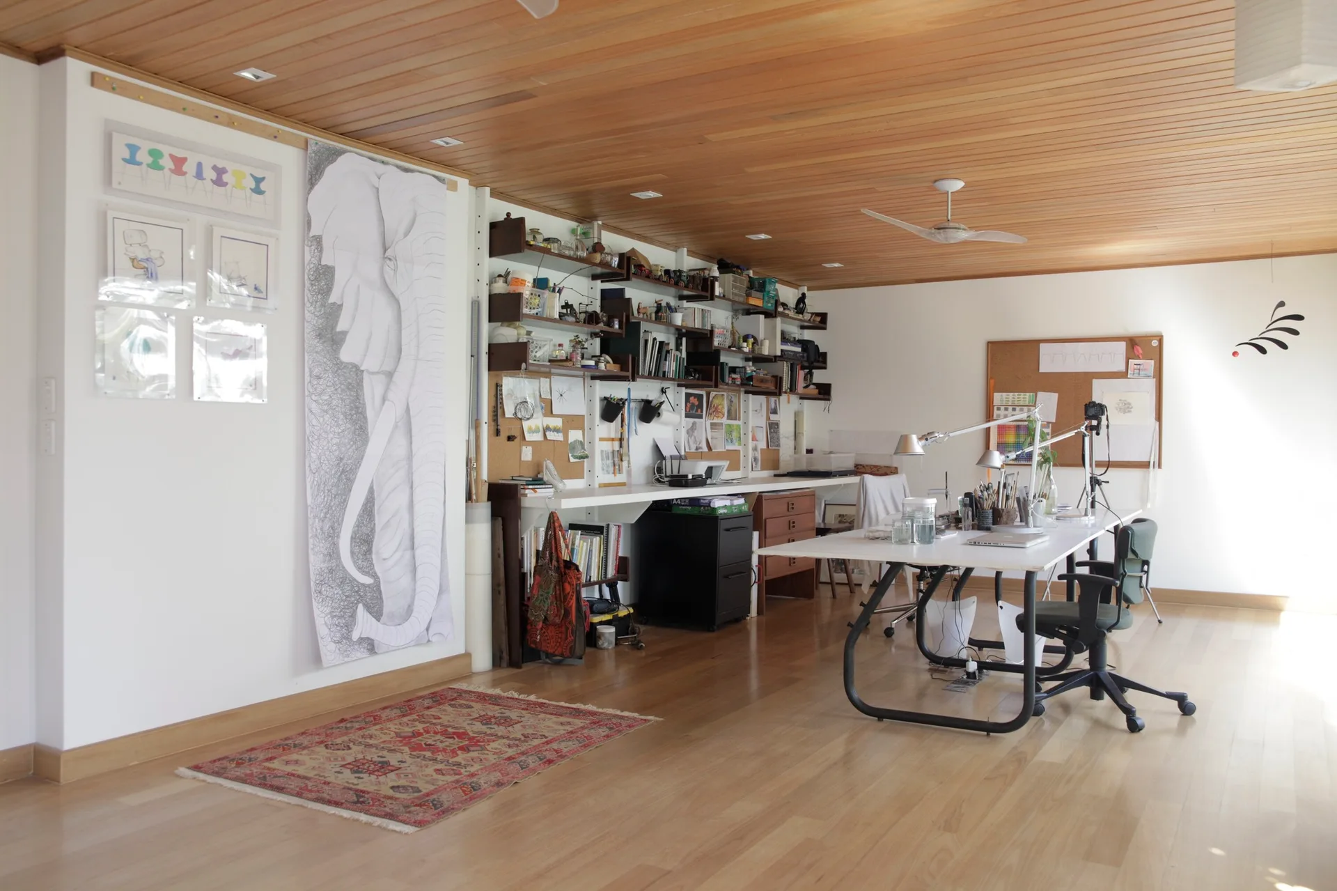 A spacious home office featuring a long white desk, adjustable task lighting, open wall shelves, and a polished wood plank ceiling.