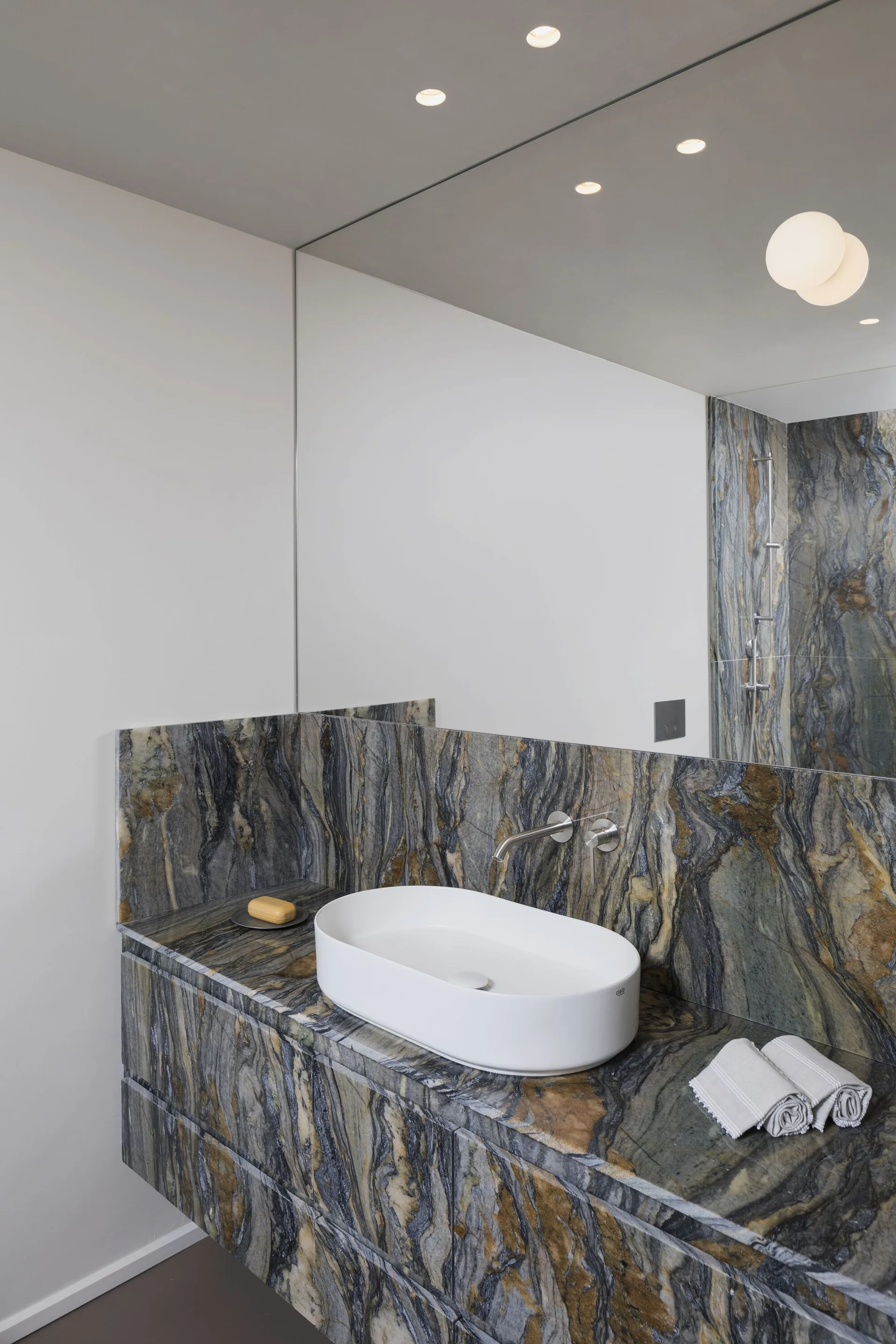 A modern floating bathroom vanity wrapped in highly veined stone features a white ceramic vessel sink and a wall-mounted faucet.