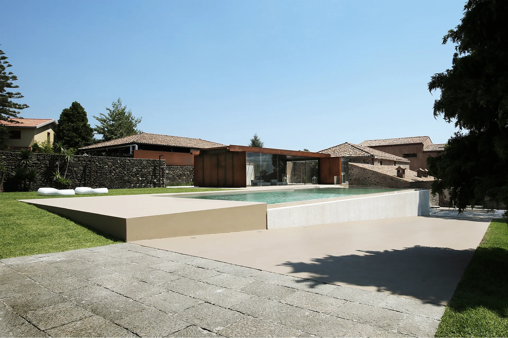 A rectangular swimming pool is surrounded by a smooth beige deck, manicured grass, and a modern building with a rust-toned facade.