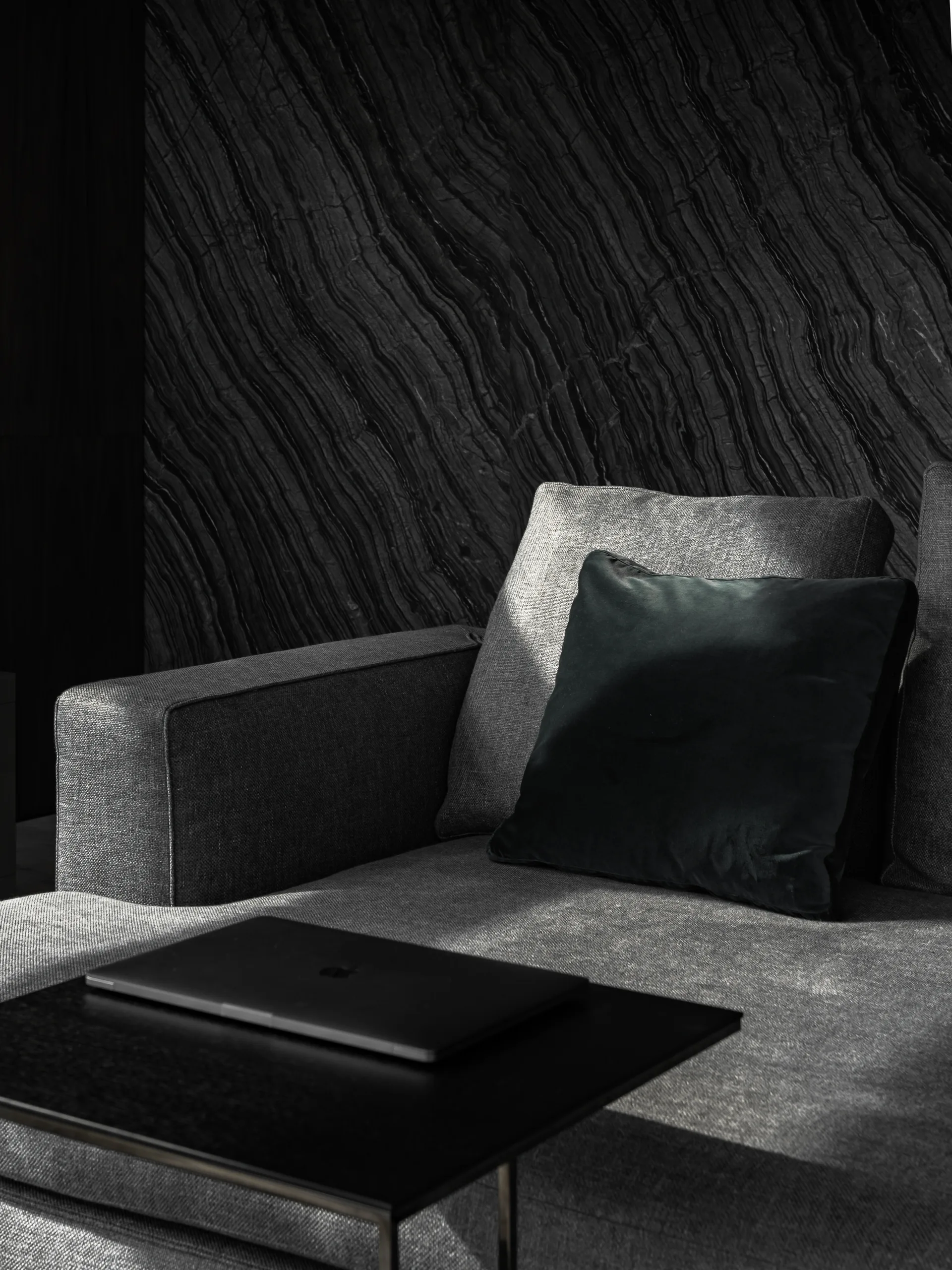 A charcoal grey upholstered sofa with a dark velvet throw pillow is positioned in front of a textured black wall panel with diagonal stripes.