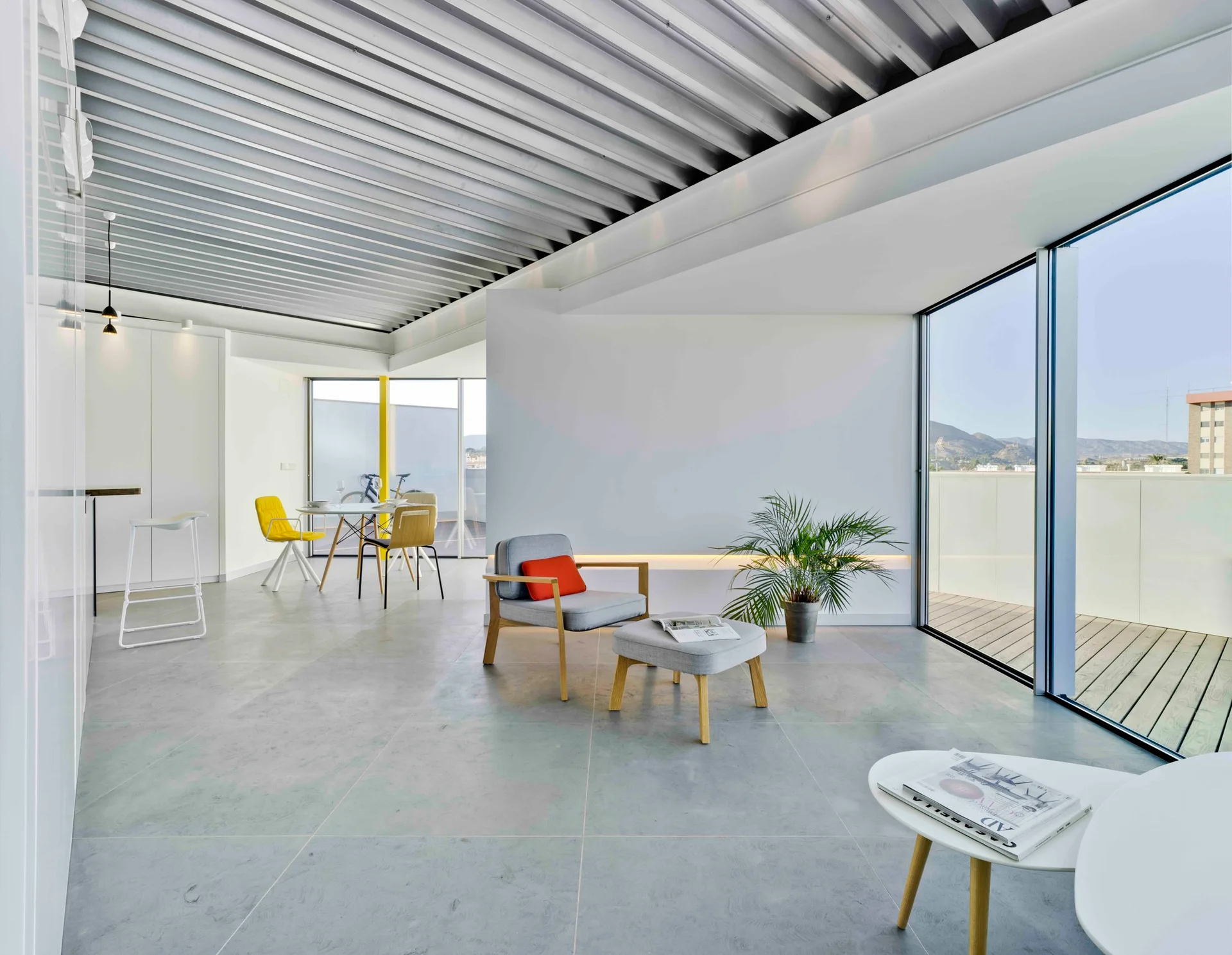 A modern open-concept living area features a grey tiled floor, a ribbed metal ceiling, a minimalist armchair, and large glass doors leading to a wood terrace.
