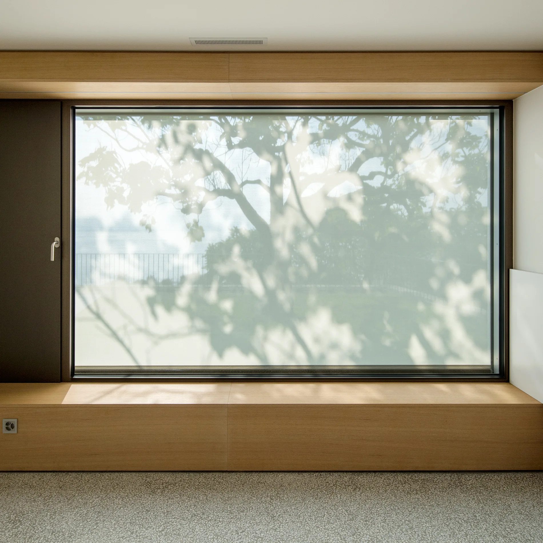 A large frosted glass window with tree shadows is framed by light wood panels and a low built-in bench above a terrazzo floor.