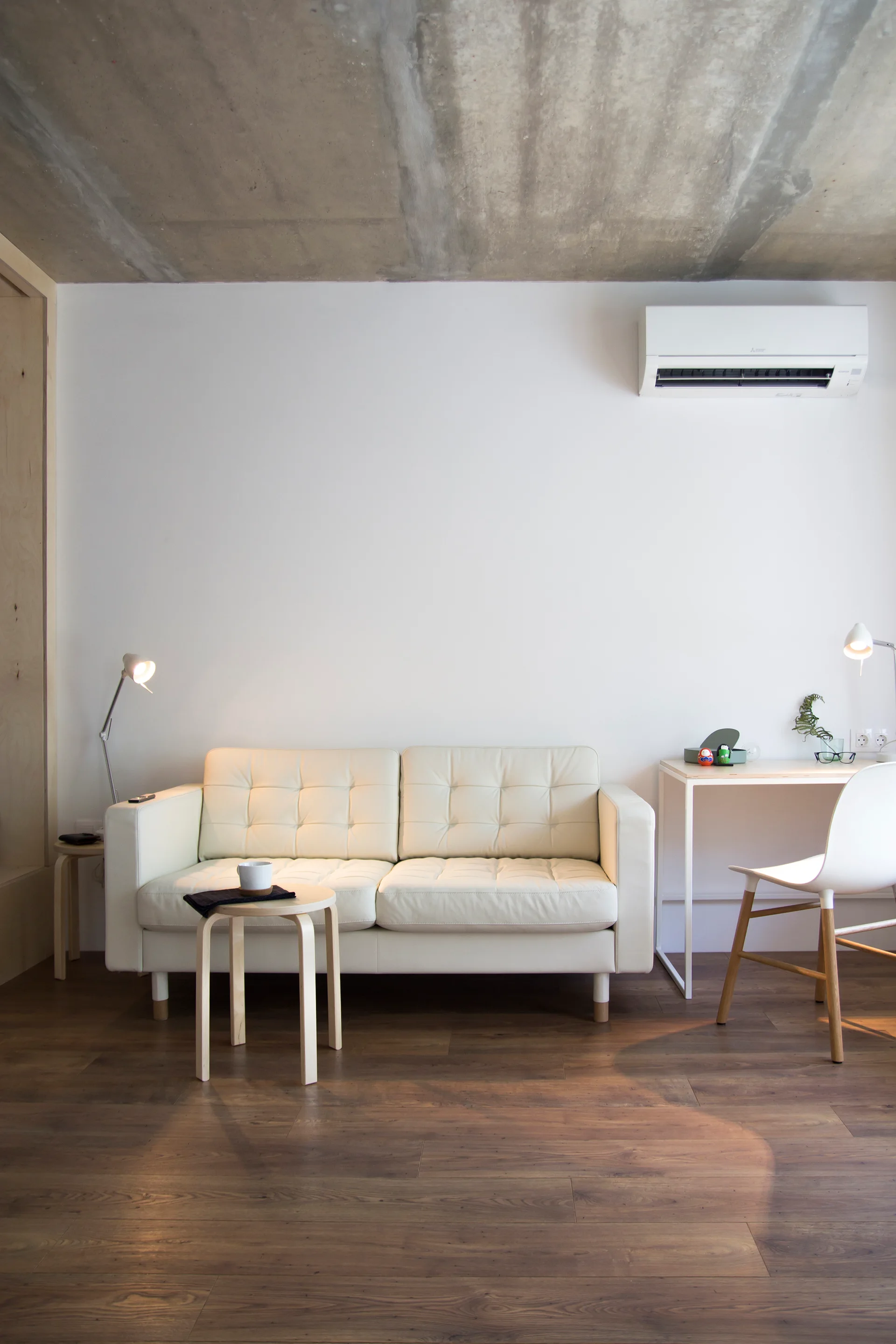 A white tufted leather sofa sits beneath an exposed concrete ceiling, flanked by a small desk and a light wood stool on dark hardwood floors.