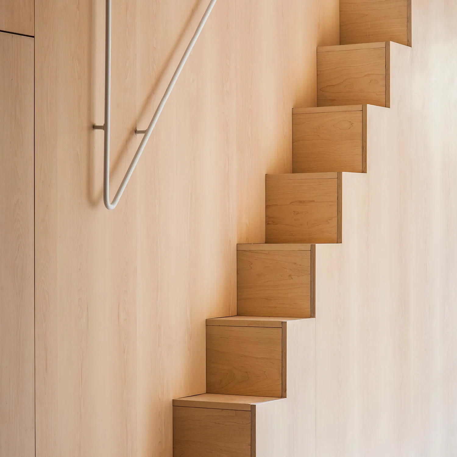 A minimalist light wood staircase with boxy steps built flush against a matching wood paneled wall next to a white handrail.