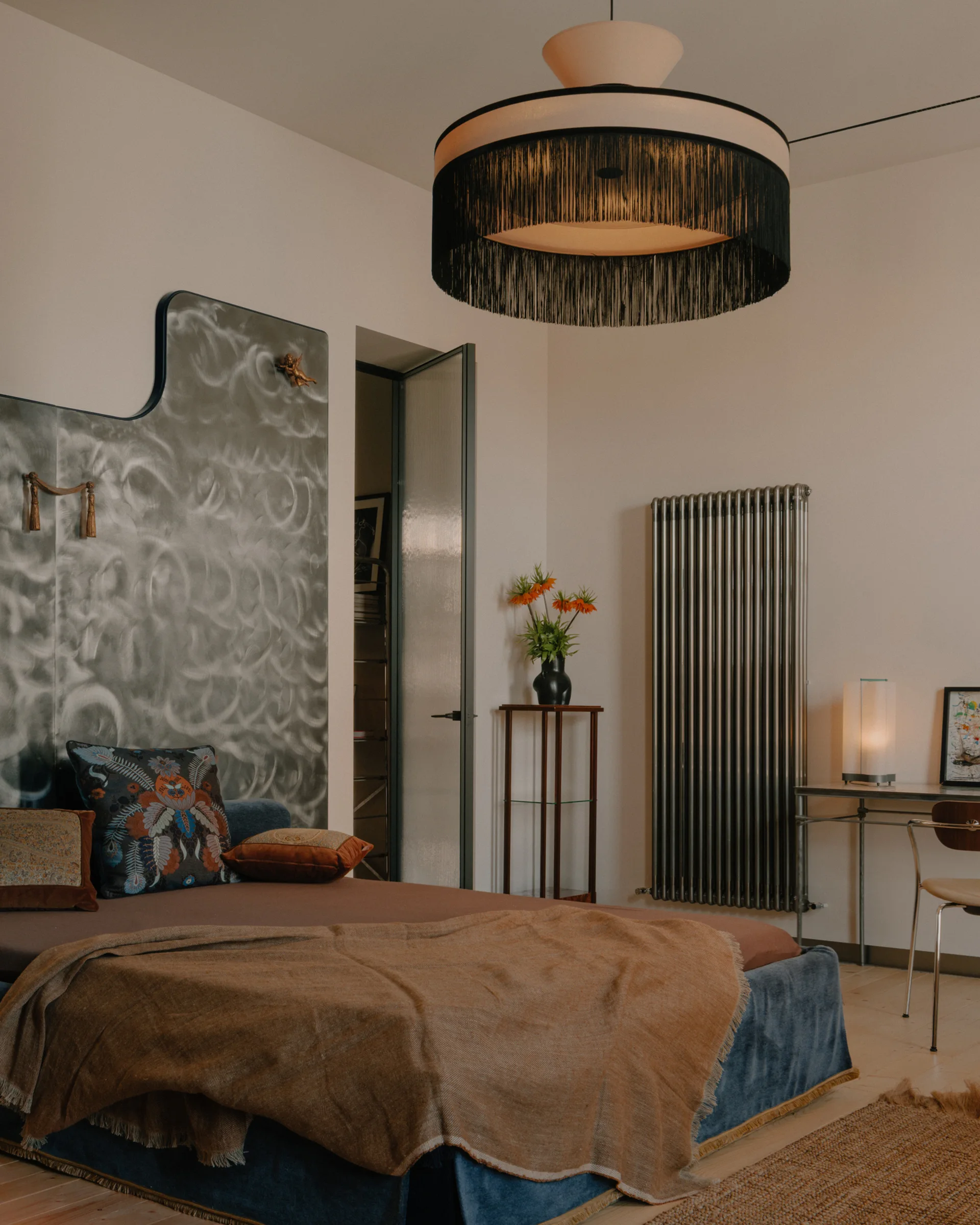 An eclectic bedroom featuring a blue upholstered bed, a textured silver wall panel, a tall column radiator, and a large black fringe pendant light.