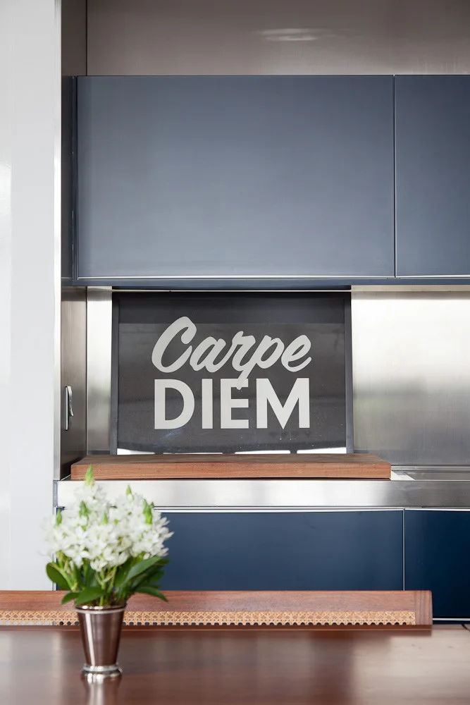 Matte blue kitchen cabinets and a stainless steel backsplash are shown behind a wooden dining table topped with a silver vase of white flowers.