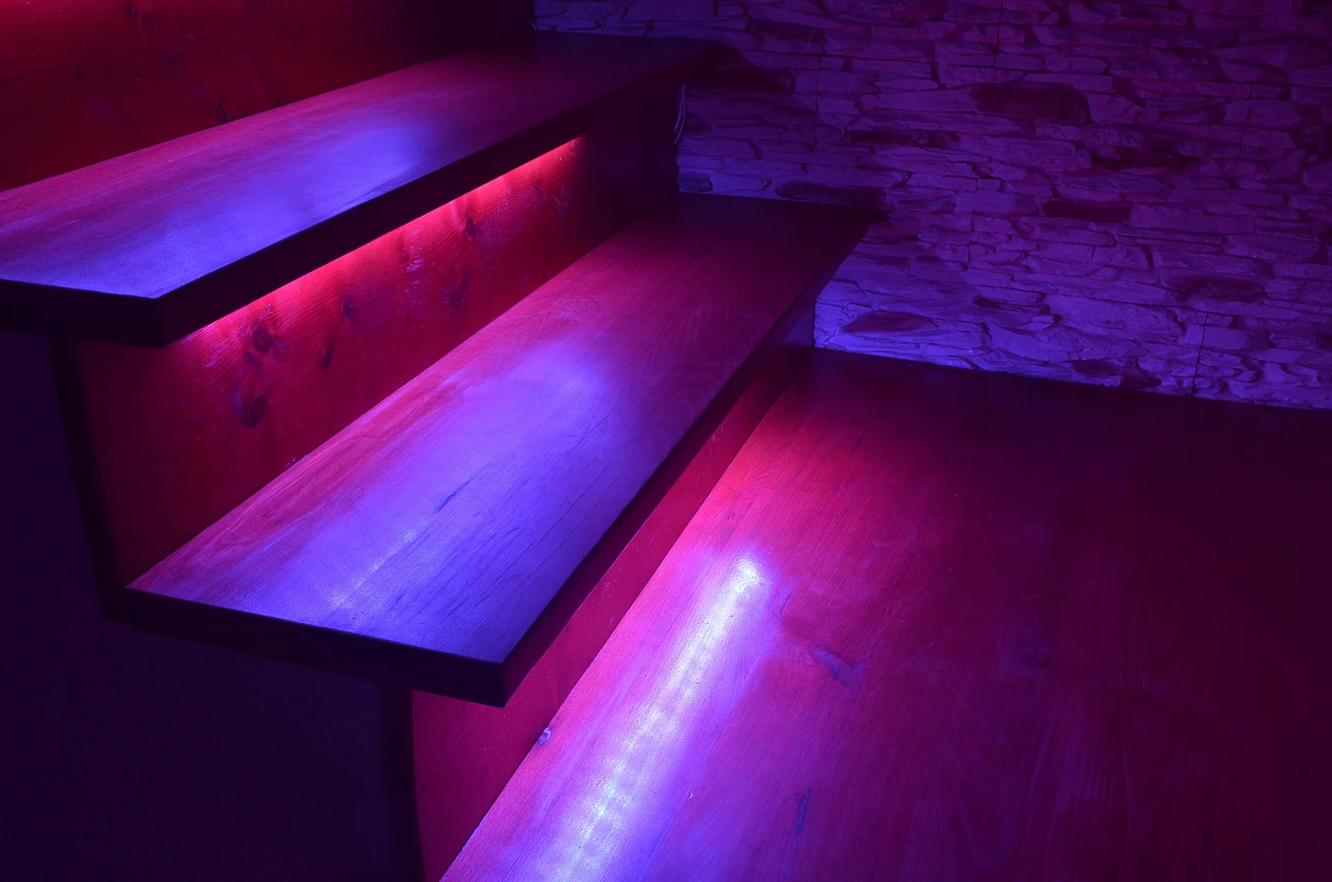 Dark wood staircase steps lit by purple and blue LED strip lights next to a rough stone accent wall.