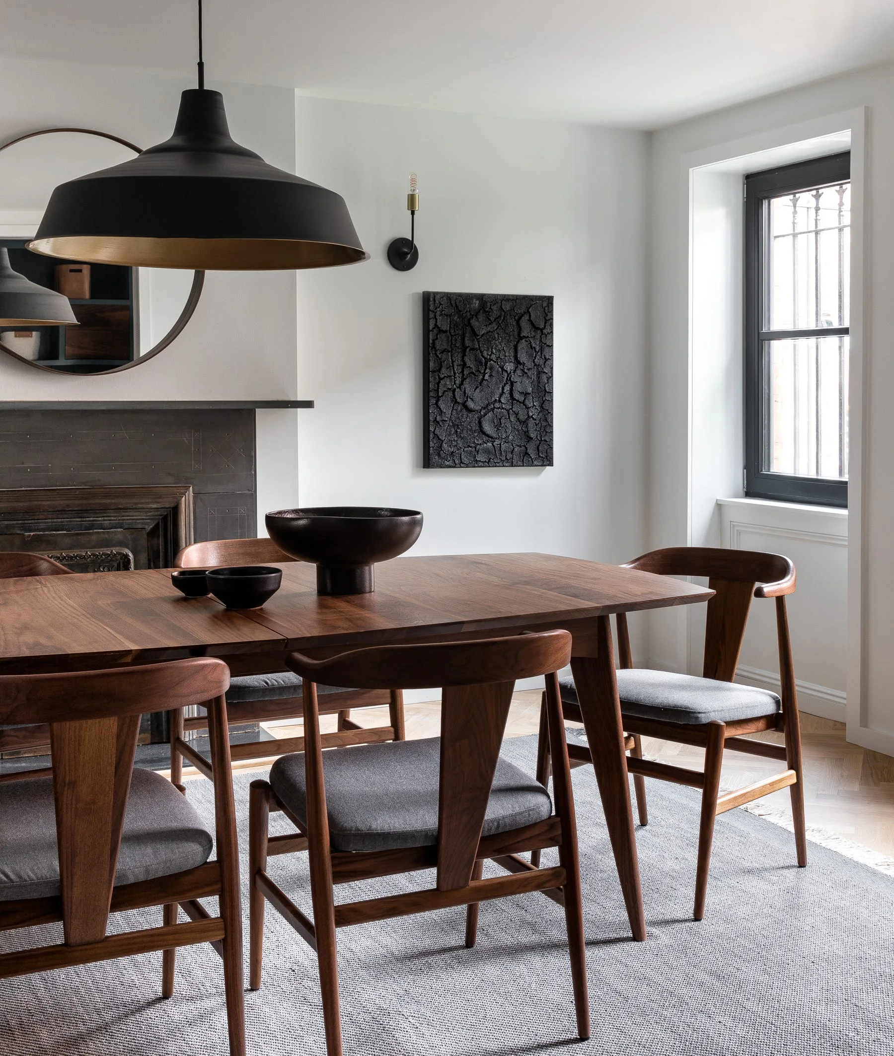 A wooden dining table and matching chairs sit on a grey rug under a large black dome pendant light in a room with white walls.