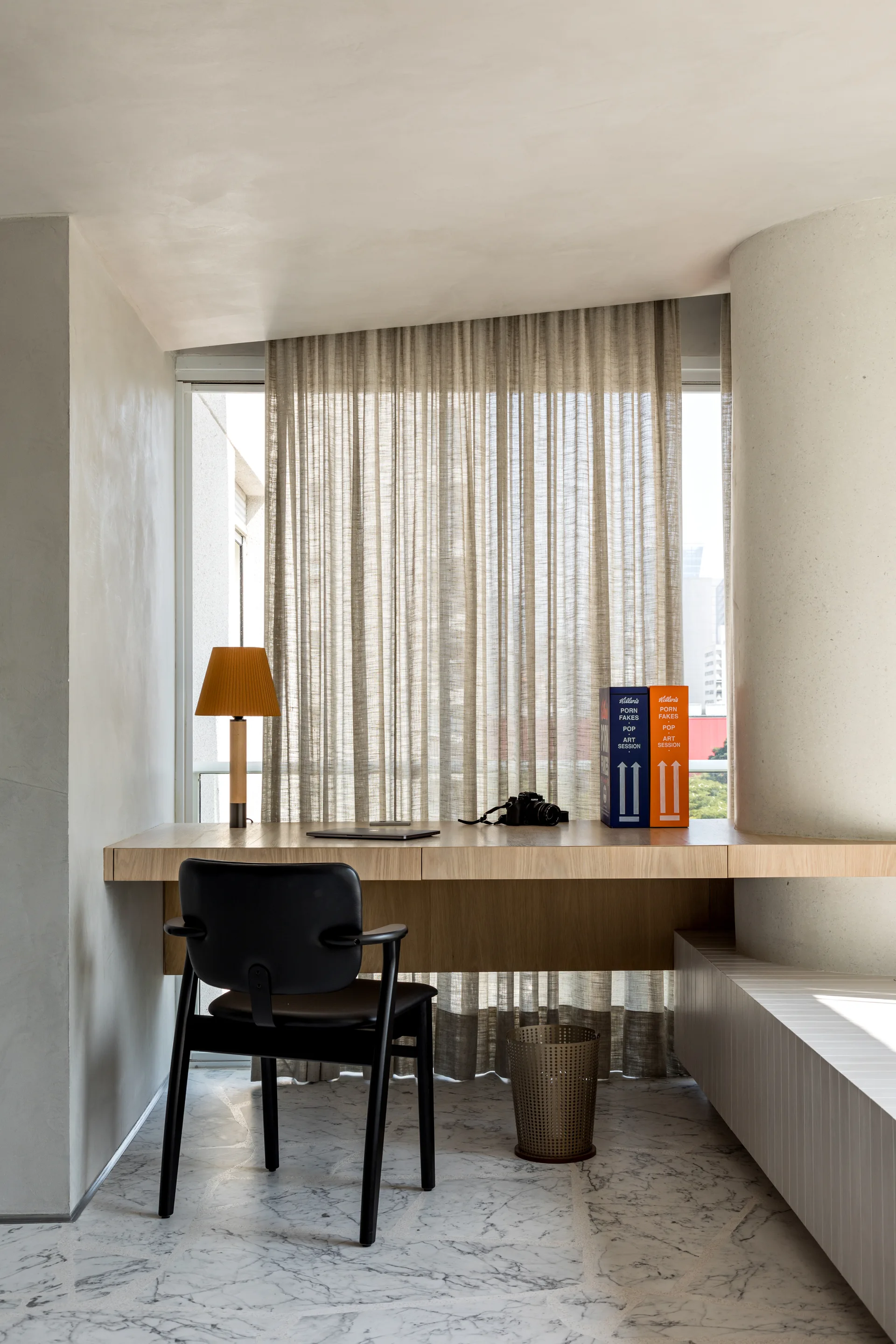 A minimalist home office features a cantilevered wood desk, a black chair, and sheer beige curtains over a white veined marble floor.