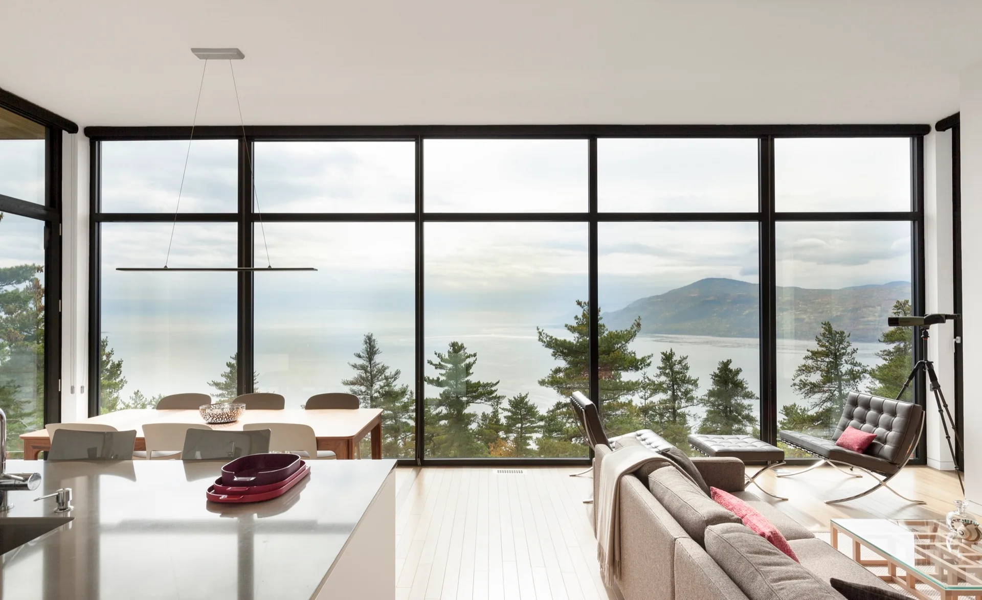 A modern open-plan living and dining area features floor-to-ceiling windows overlooking a lake and mountains, a grey sofa, and a wooden table.