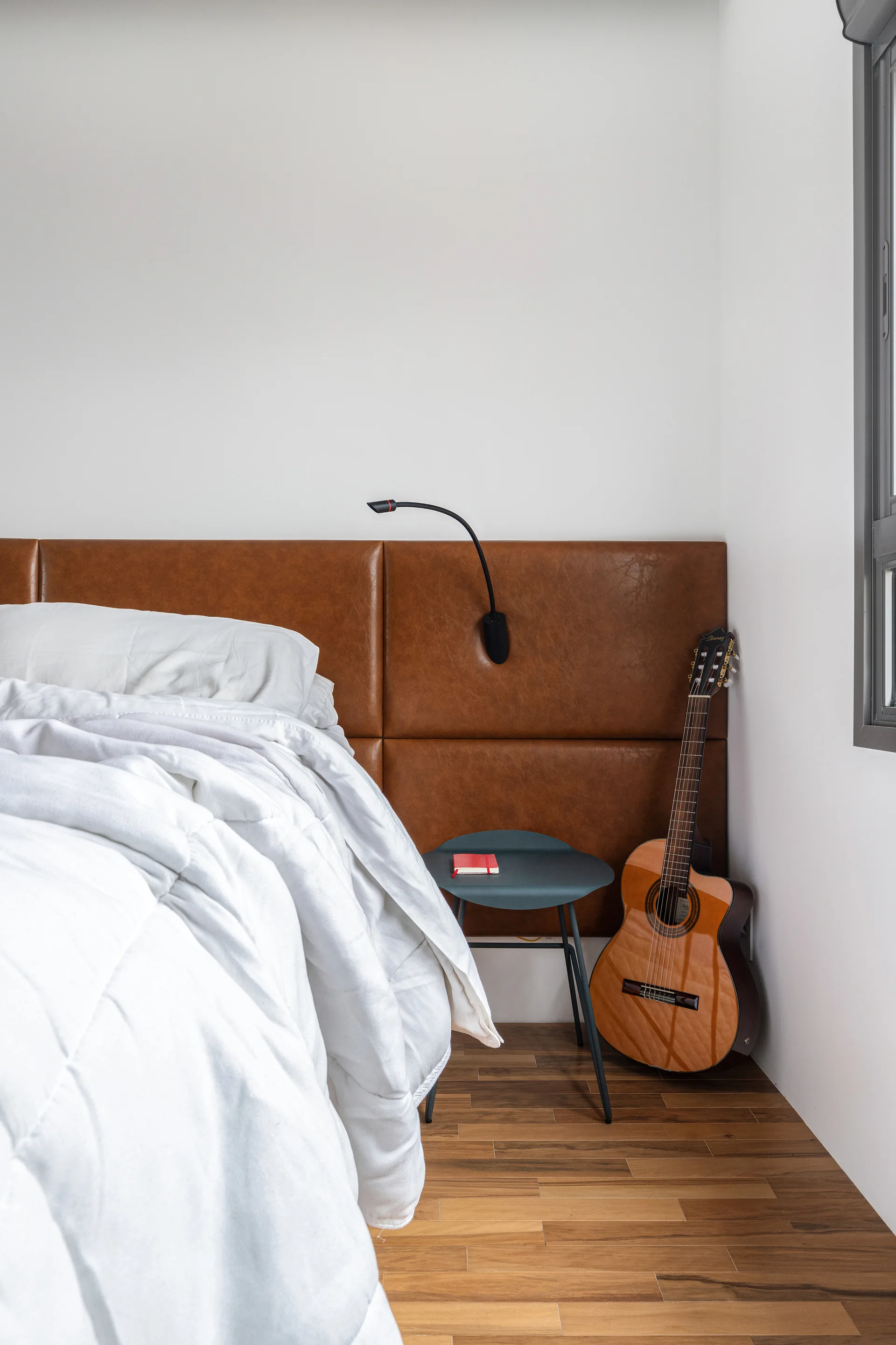 A bedroom with a brown leather paneled headboard, white bedding, a teal bedside table, and an acoustic guitar leaning against the white wall.