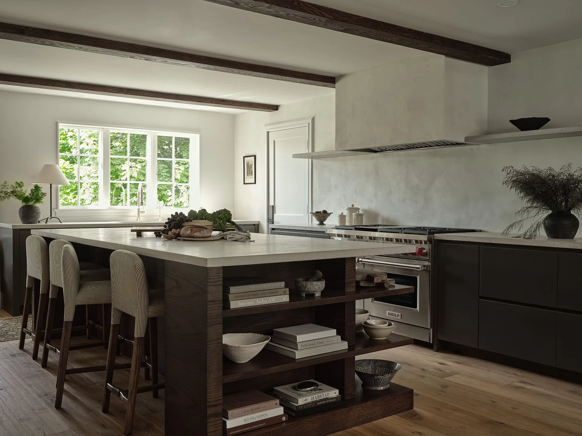 A modern rustic kitchen featuring a dark wood island with open shelving and striped bar stools underneath exposed timber ceiling beams.