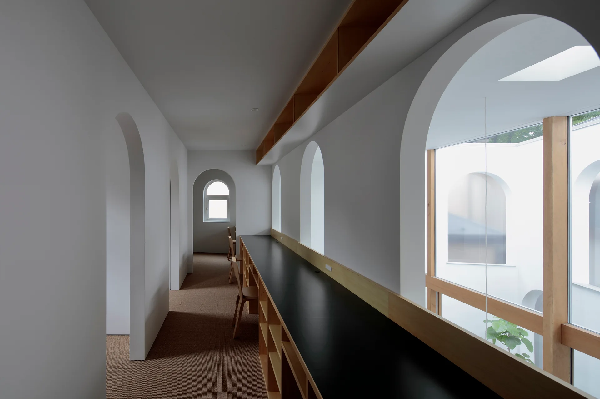 A minimalist white hallway features a long black built-in desk with wooden chairs, set alongside multiple arched window openings.