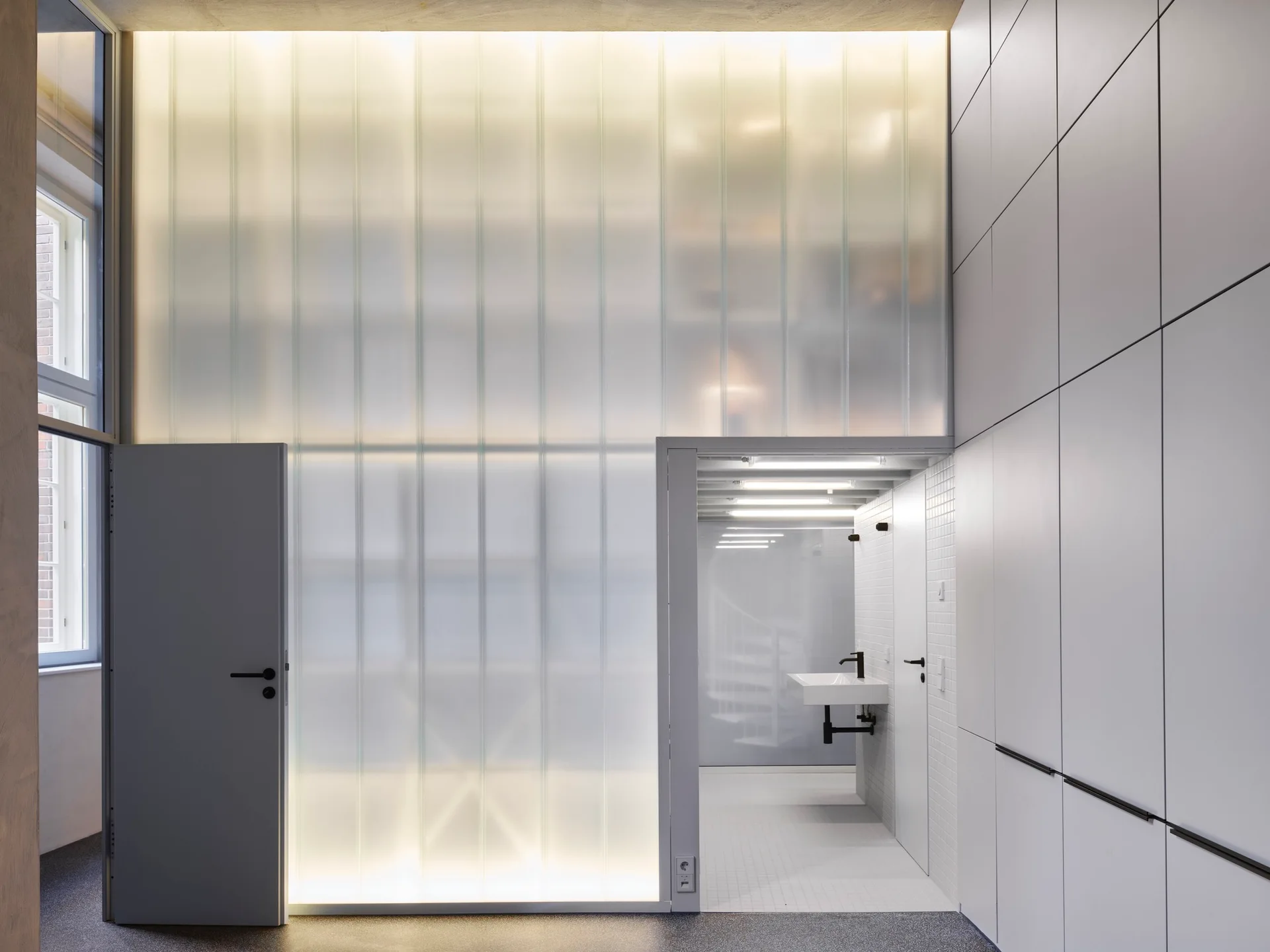A minimalist bathroom area featuring a backlit translucent glass wall partition, a wall-mounted sink, white tiled walls, and gray storage cabinets.
