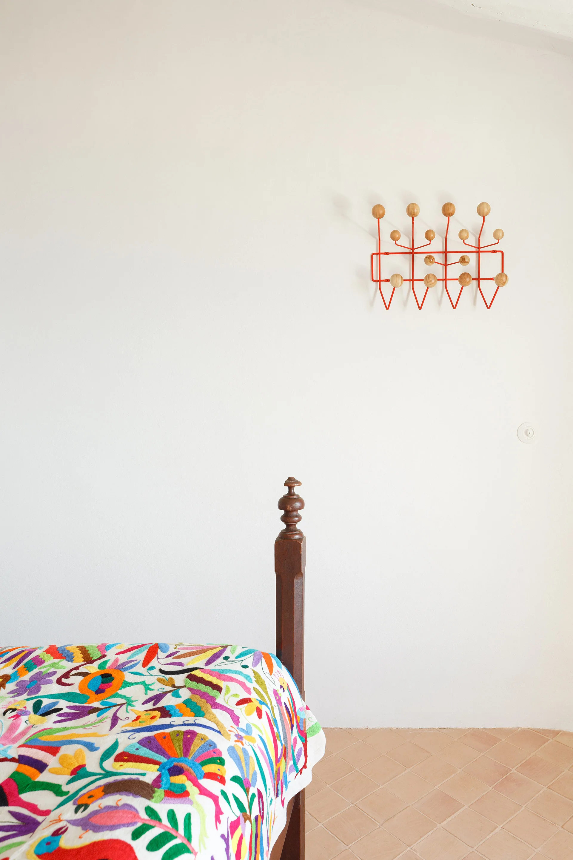 A minimalist bedroom featuring a wooden bedpost with a colorful embroidered bedspread, a red wire coat rack on a white wall, and terracotta tile floors.