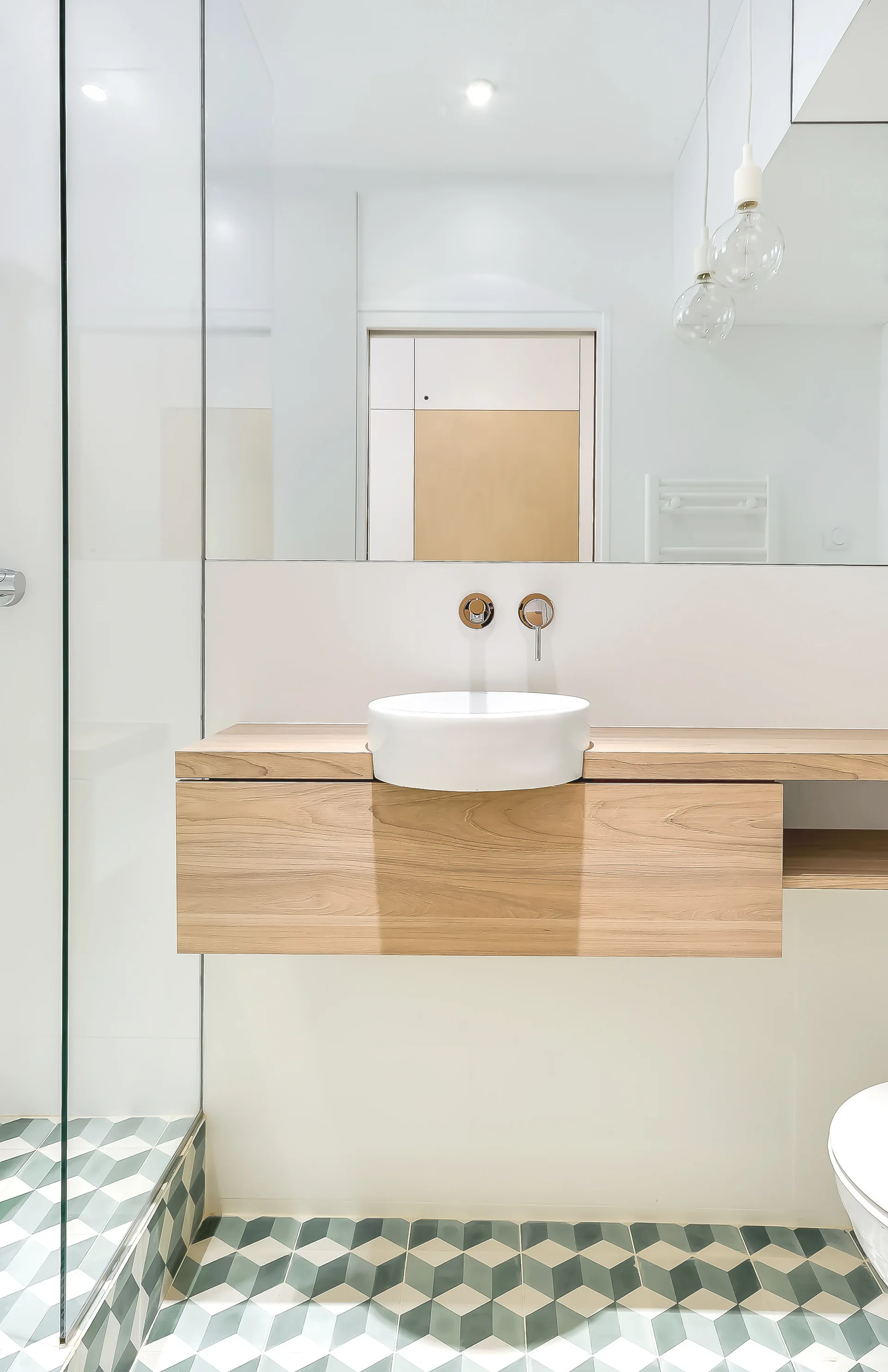 A modern bathroom features a floating wood vanity with a white vessel sink, a large mirror, and green and white geometric floor tiles.