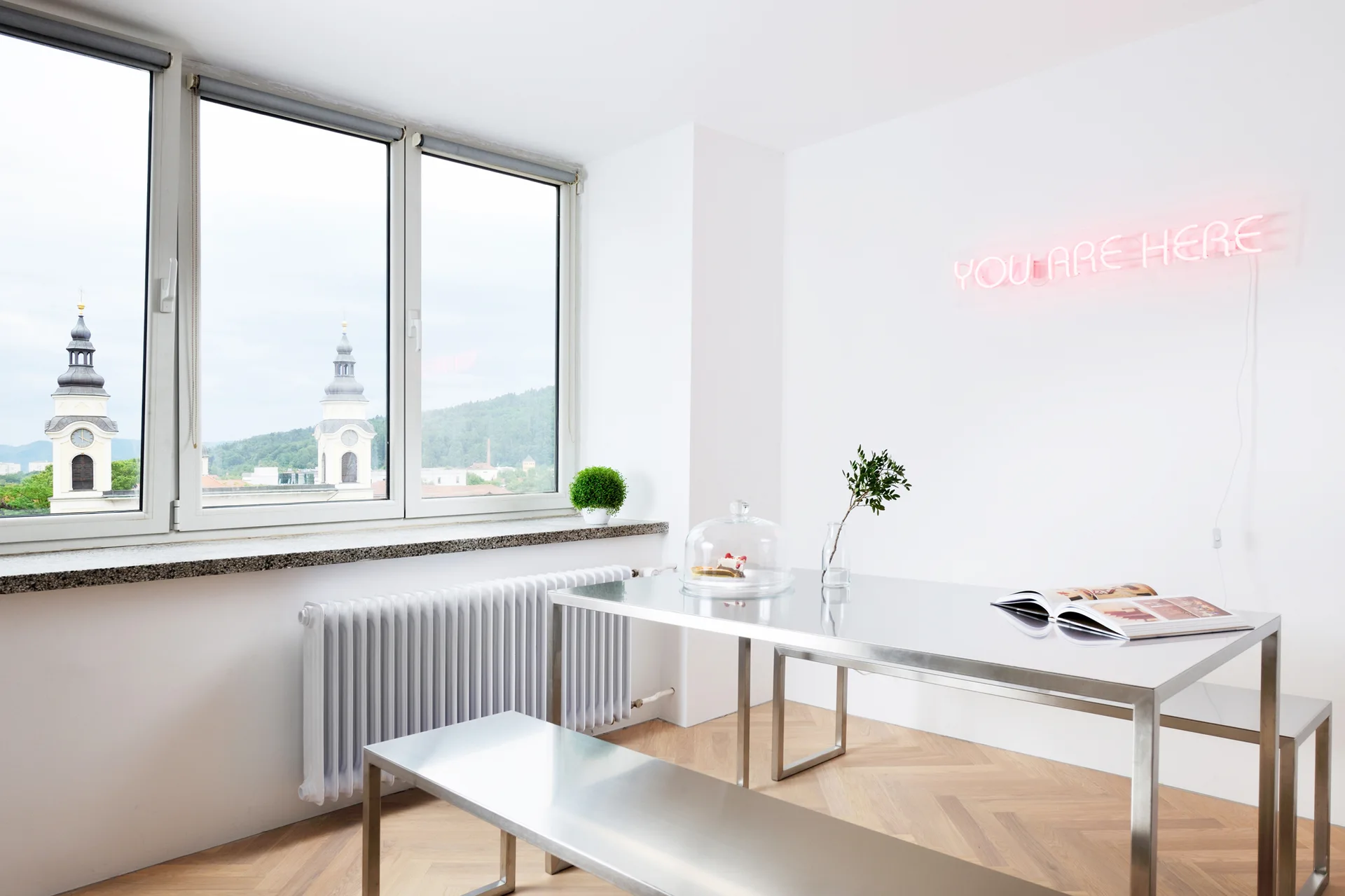 A minimalist dining room features a stainless steel table and benches on a herringbone wood floor next to a window and a pink neon wall sign.