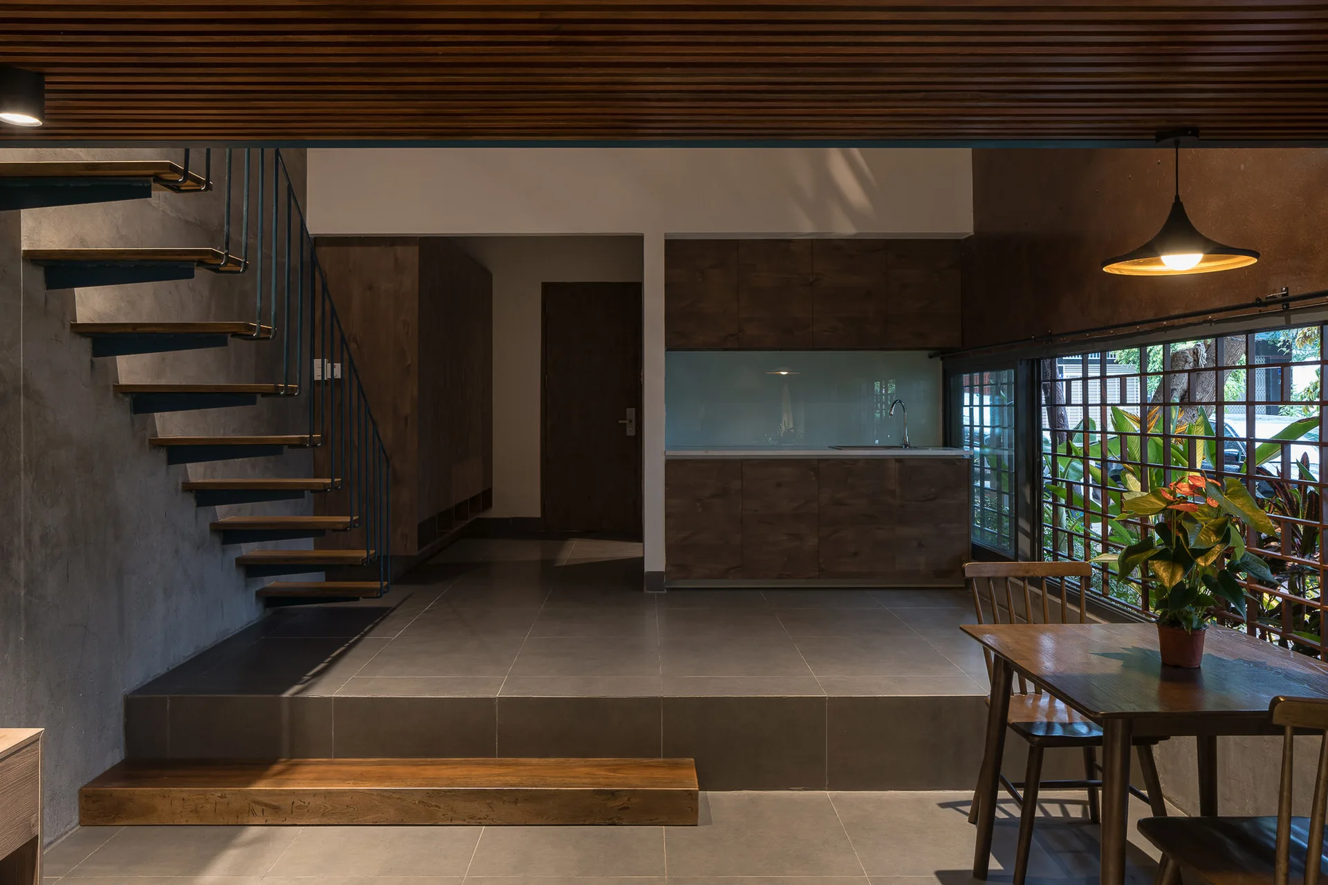 A modern split-level interior featuring an open-riser wooden staircase, dark cabinetry, and a dining table under a wooden slat ceiling.