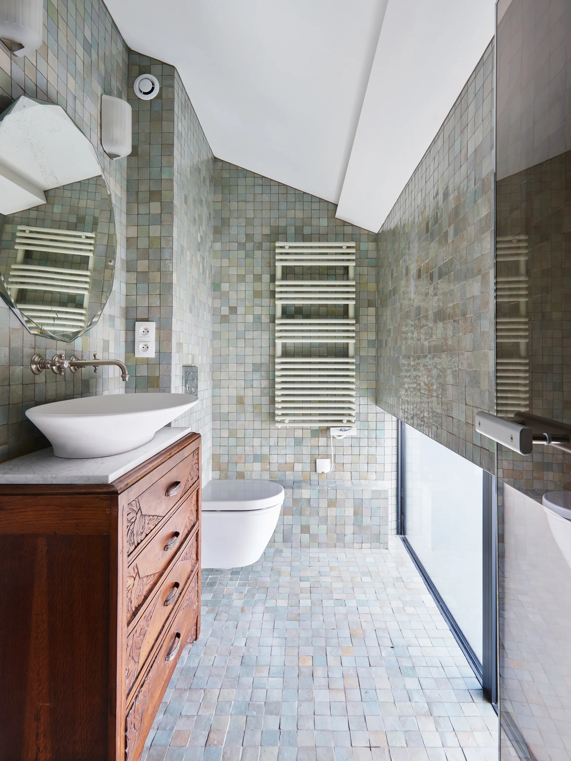 A compact bathroom tiled in green and grey squares features a carved wooden vanity with a white vessel sink and a wall-mounted toilet.