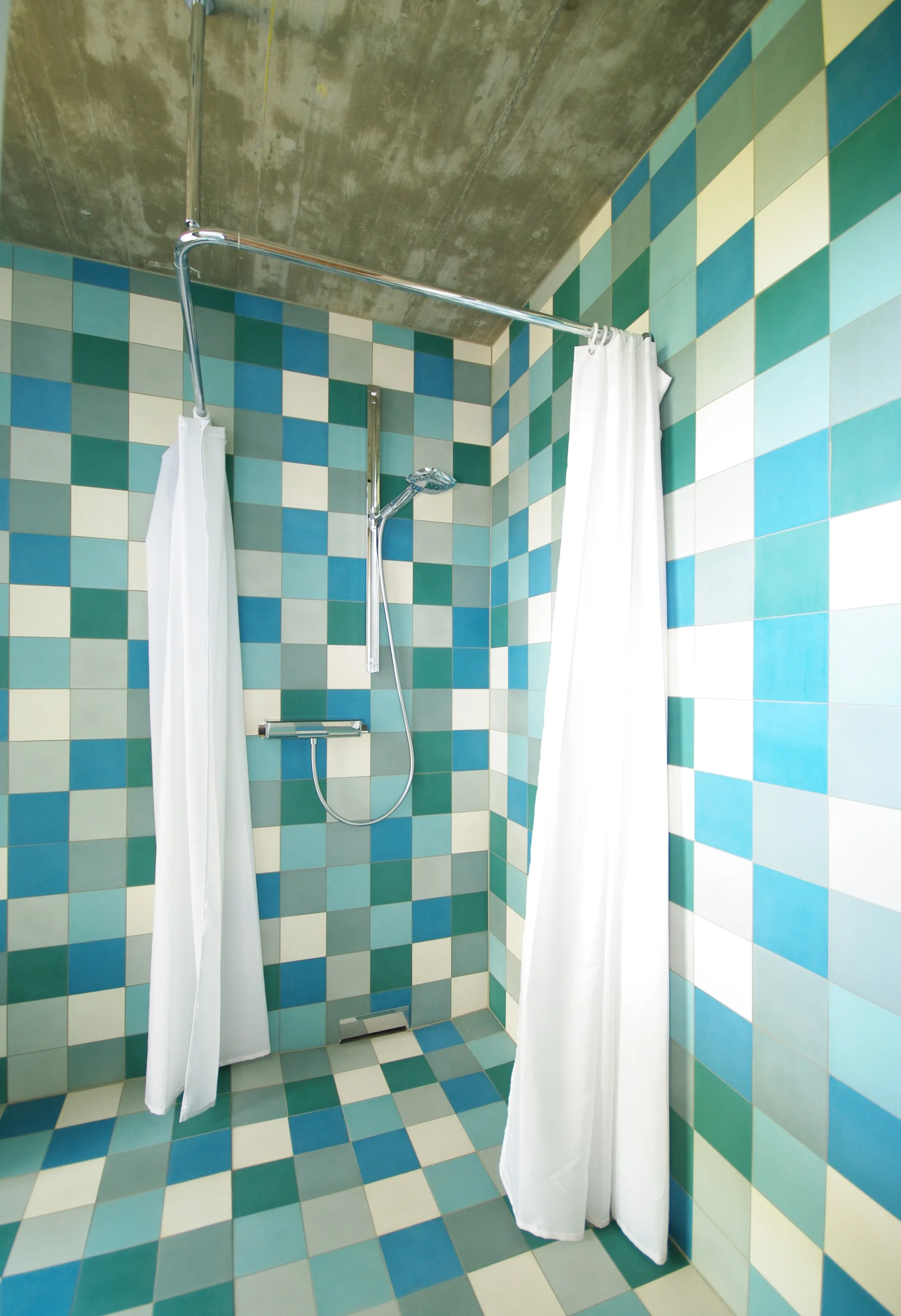 A corner shower stall is covered in a grid pattern of blue, teal, and white square tiles beneath an exposed concrete ceiling.