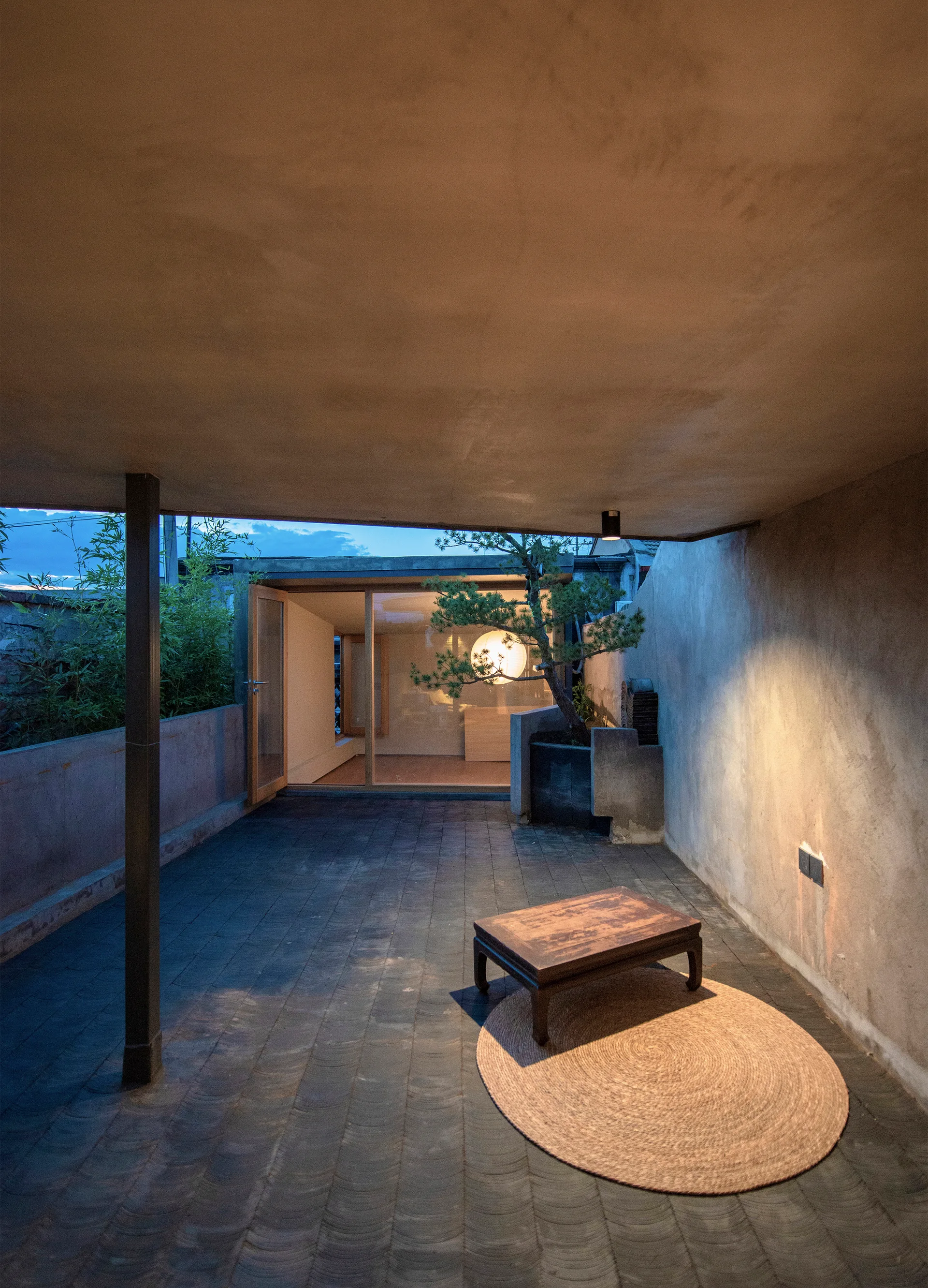 A covered concrete patio features a low wooden table on a round woven rug next to a potted bonsai tree under dim evening light.