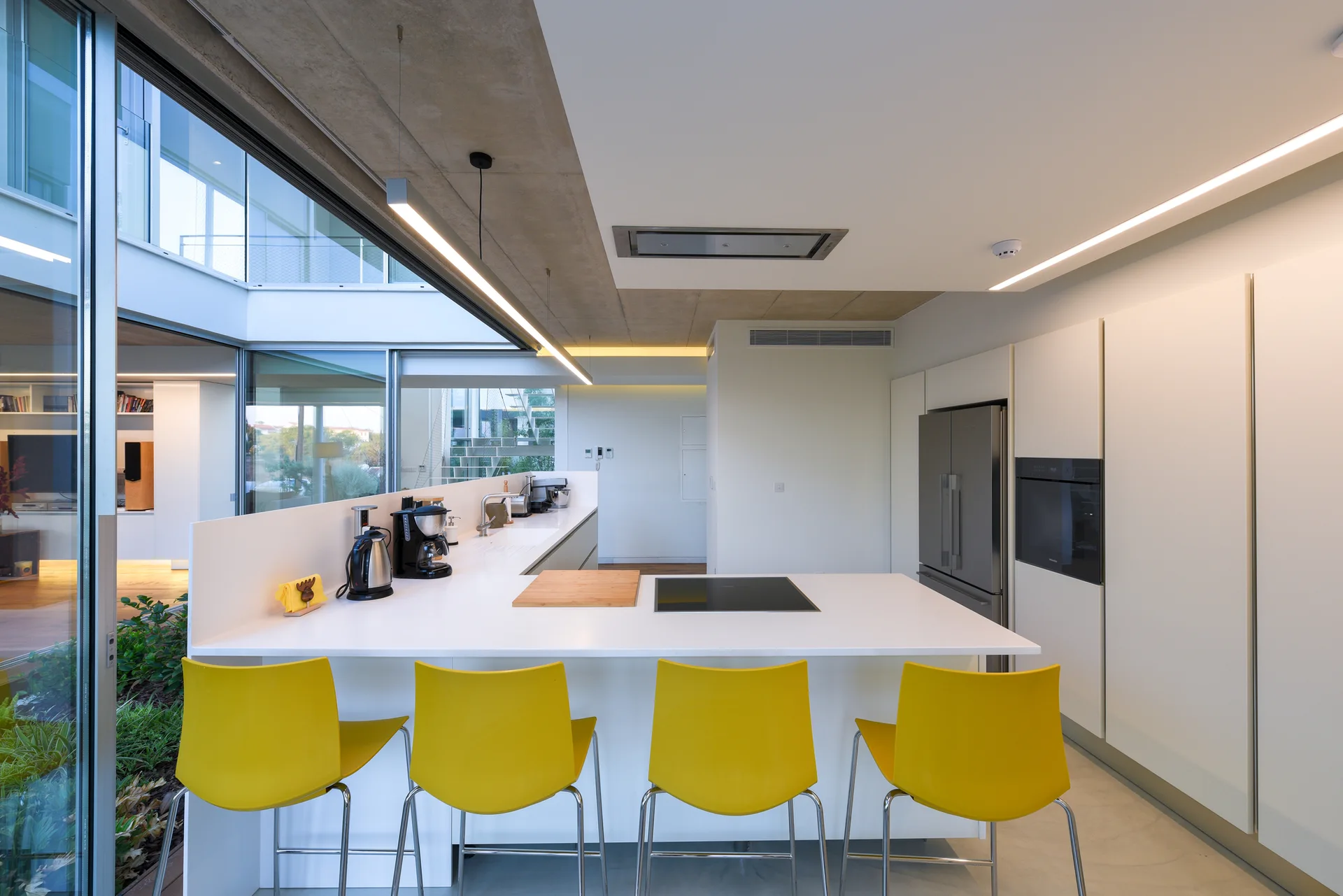 Four bright yellow bar stools sit at a white minimalist kitchen island next to floor-to-ceiling glass windows and white seamless cabinetry.