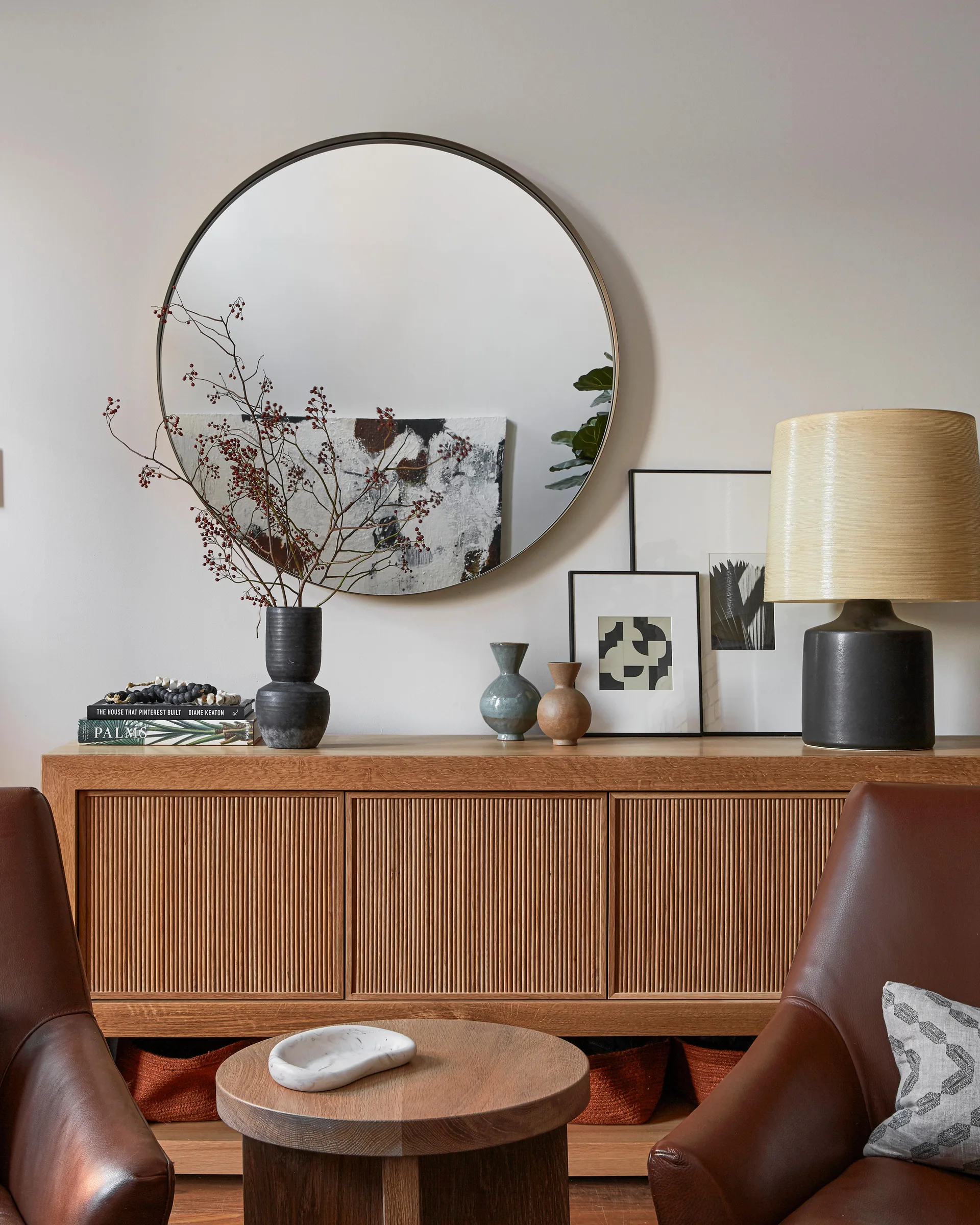 A ribbed wood console table styled with a large round mirror, abstract art, ceramic vases, and a lamp between two brown leather armchairs.
