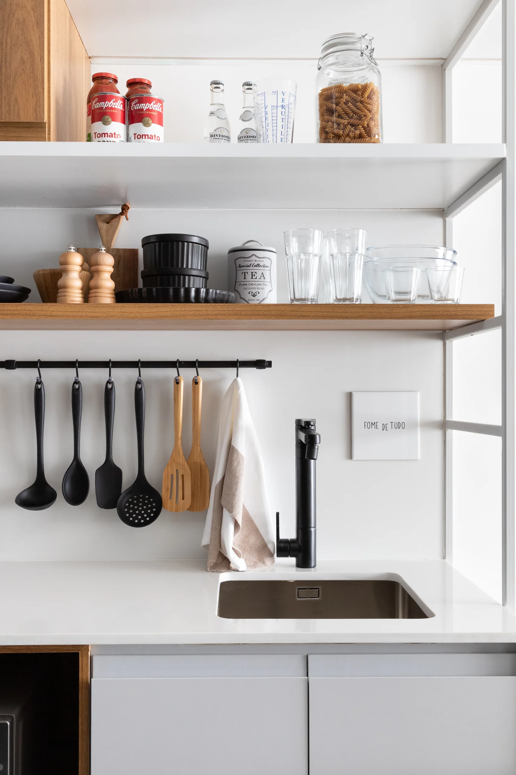 A minimalist kitchen prep area features a matte black faucet, an undermount sink, a black utensil rail, and open wood and white shelving.