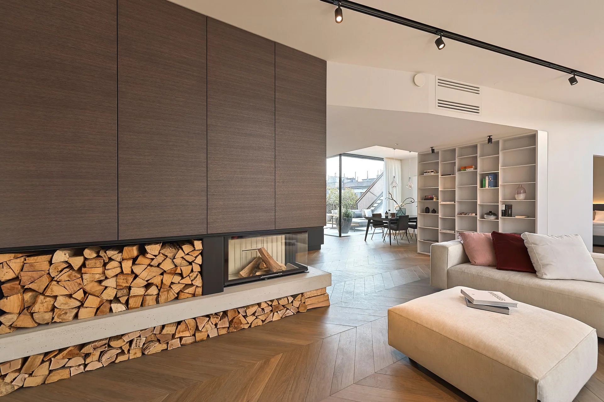 A modern living room features a built-in fireplace with stacked firewood, a beige sofa, and a brown herringbone wood floor.